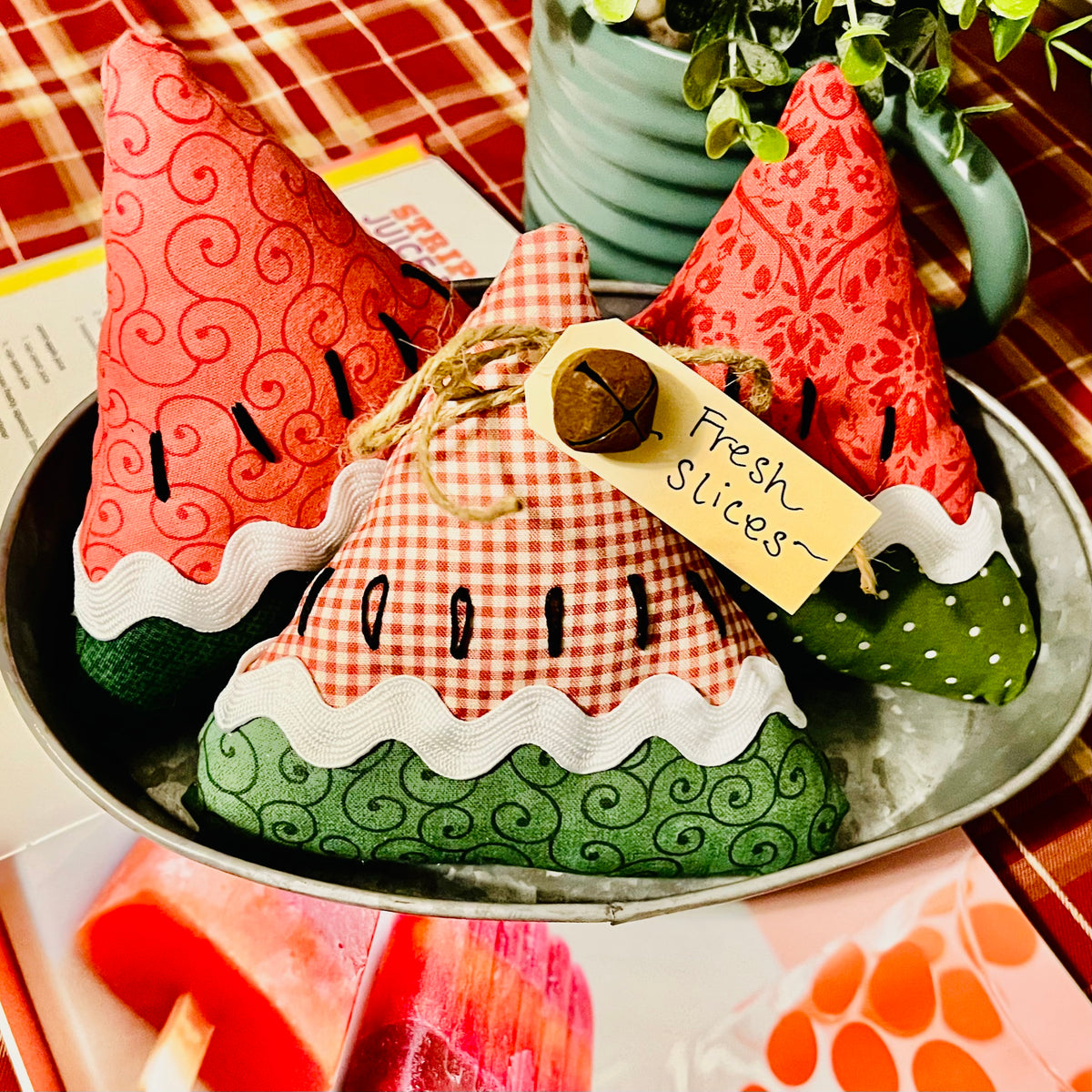 Three decorative fabric watermelon slices on a metal tray with a plant in the background.