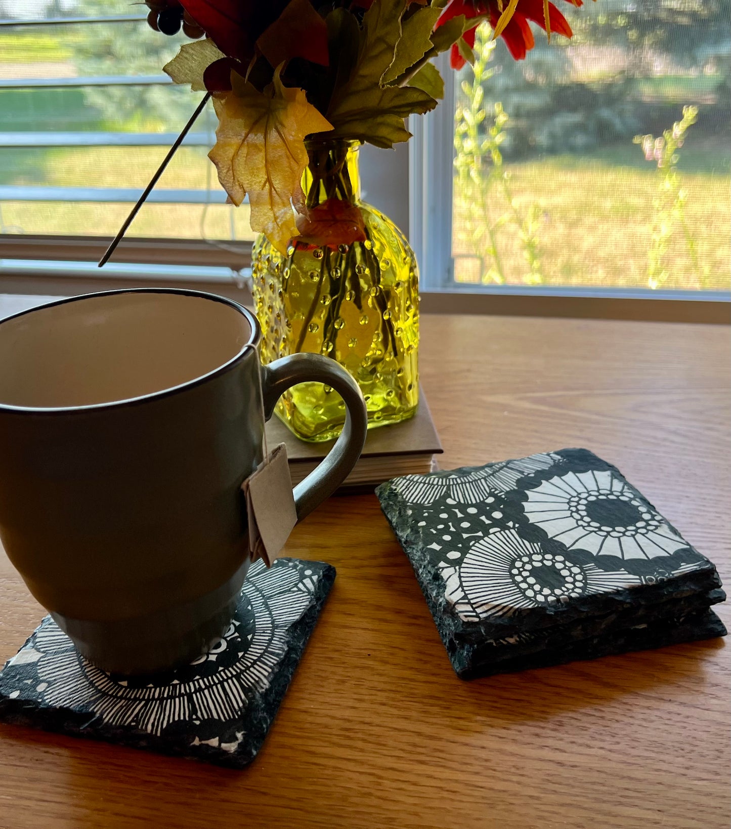 A black and white patterned slate coaster made by Eclectic Spruce sitting on a wooden table, with a gray ceramic mug resting on one coaster and a stack of matching coasters beside it. A yellow glass vase with dried flowers sits in the background near a window.