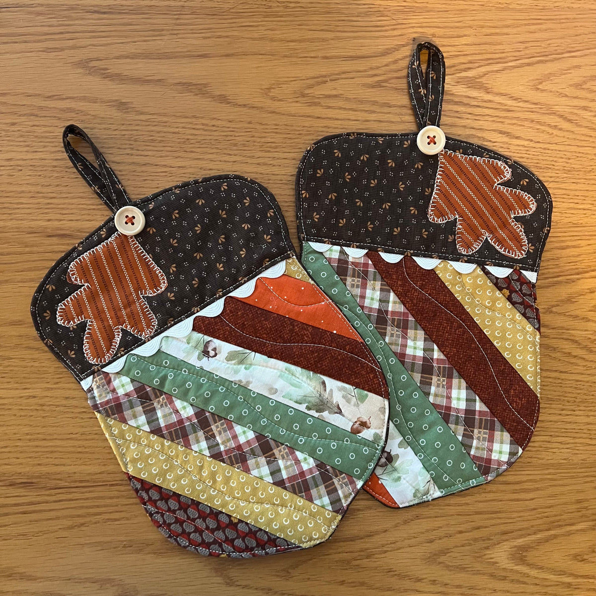 A set of two Eclectic Spruce acorn-shaped quilted potholders with autumn-themed patchwork fabrics, applique leaf design, wood buttons, rick rack detailing, and hanging loops.