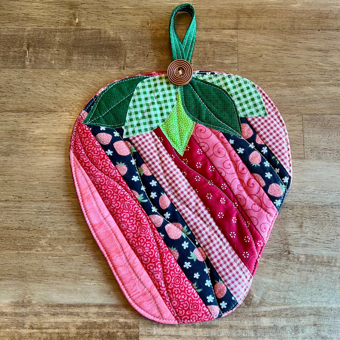 One strawberry-shaped quilted potholder on a wooden table. This is the Eclectic Spruce Sweet Strawberry hot pad, which features strip patchwork in various pink, red, white, and coral cotton prints, green leaves, and a wooden button accent.