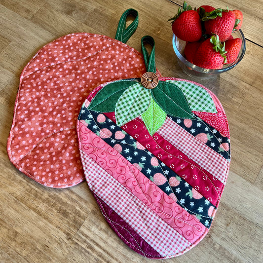 Two strawberry-shaped quilted potholders on a wooden table, with a small glass bowl of fresh strawberries in the background. One potholder is flipped upside down showing the "strawberry print" backside to the potholder. The other one features the front side of the Eclectic Spruce Sweet Strawberry hot pad, which features strip patchwork in various pink, red, white, and coral cotton prints, green leaves, and a wooden button accent.