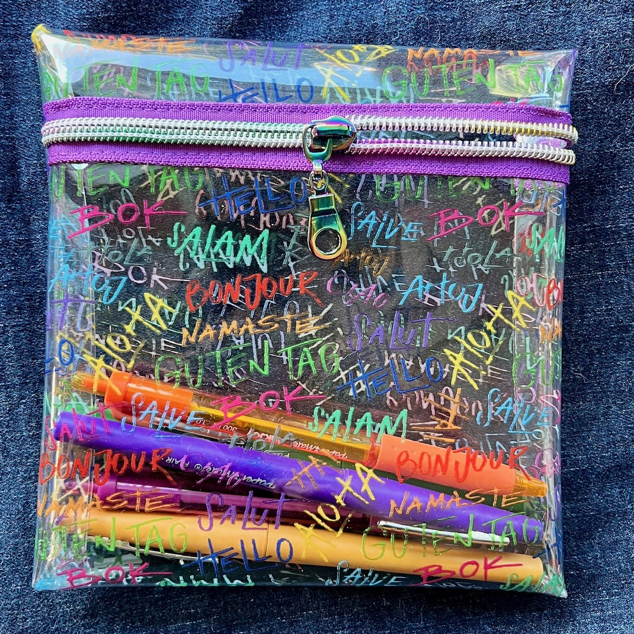 Clear see-through vinyl zipper pouch that says "hello" in various languages and filled with colorful pens on a blue denim background.