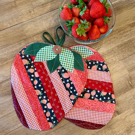 A pair of Eclectic Spruce's Sweet Strawberry Hot Pads sitting on a wooden table next to a bowl full of strawberries.