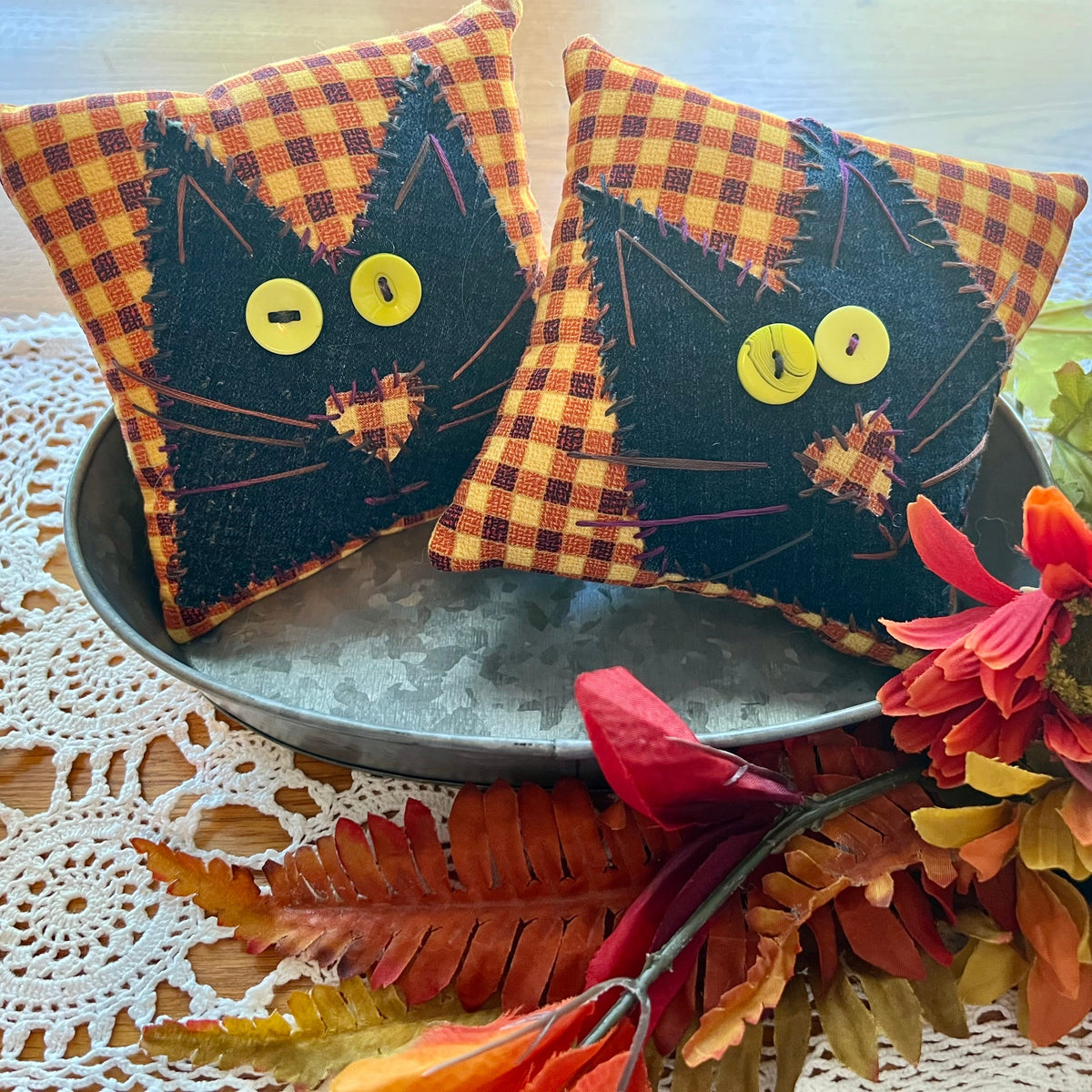 Example of two handmade Eclectic Spruce Purrfect Patchwork Cat Pillows. These are 5x5 inch decorative square pillows with black cat faces and orange plaid background, displayed in a metal tray with autumn leaves and flowers on a lace doily.