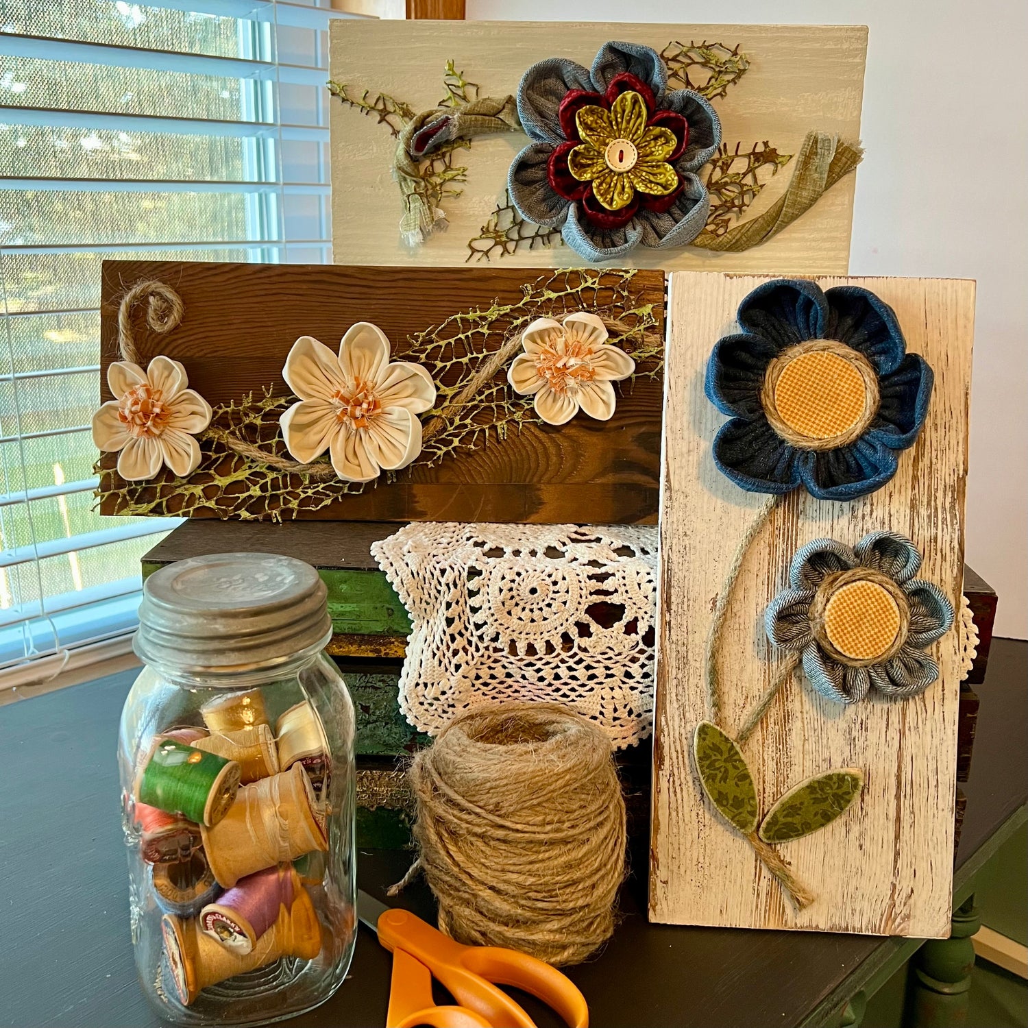 Handcrafted 3D botanical wall art featuring textile flowers mounted onto wooden planks. Three varied pieces are displayed next to a mason jar full of spools of thread, a spool of twine, and a pair of scissors.