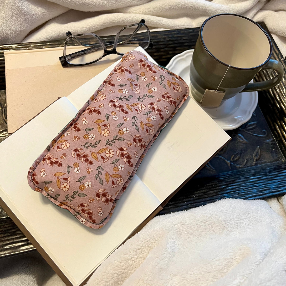 An Evening Primrose Weighted Eye Pillow by Eclectic Spruce is sitting on a tray with a book, mug, pair of glasses, and a cozy white blanket.