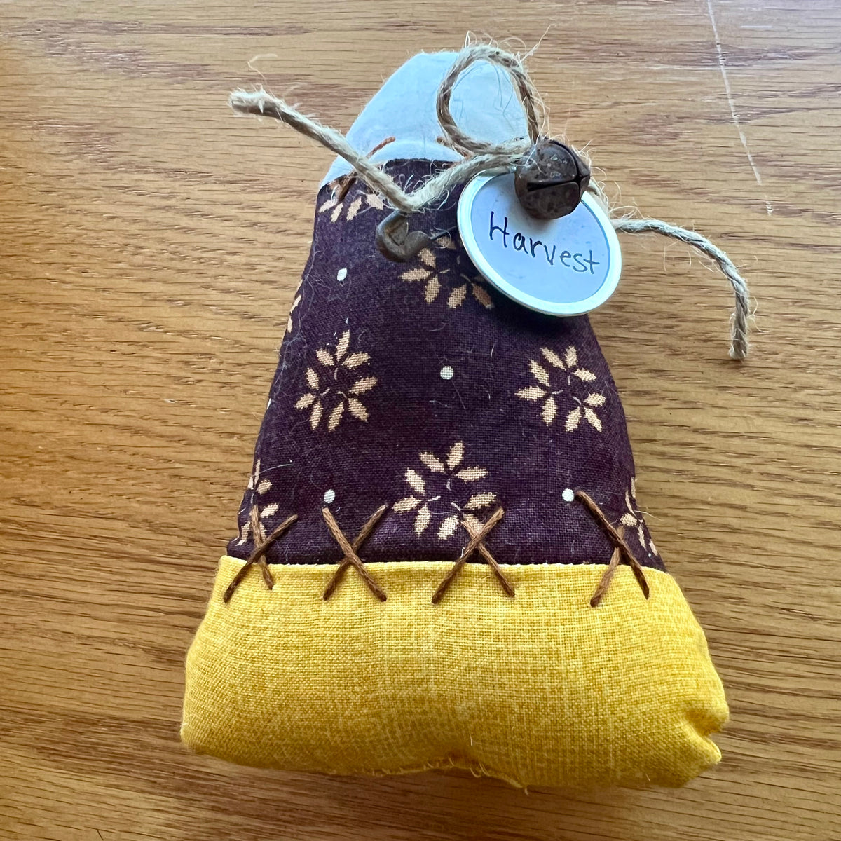 An Eclectic Spruce handmade fabric candy corn, featuring a dark brown fabric with a sunflower pattern in the middle, a mustard yellow lower section, and a white top section. It is decorated with hand stitch details, twine, a small rusty bell, and a round tag that reads 'Harvest'.