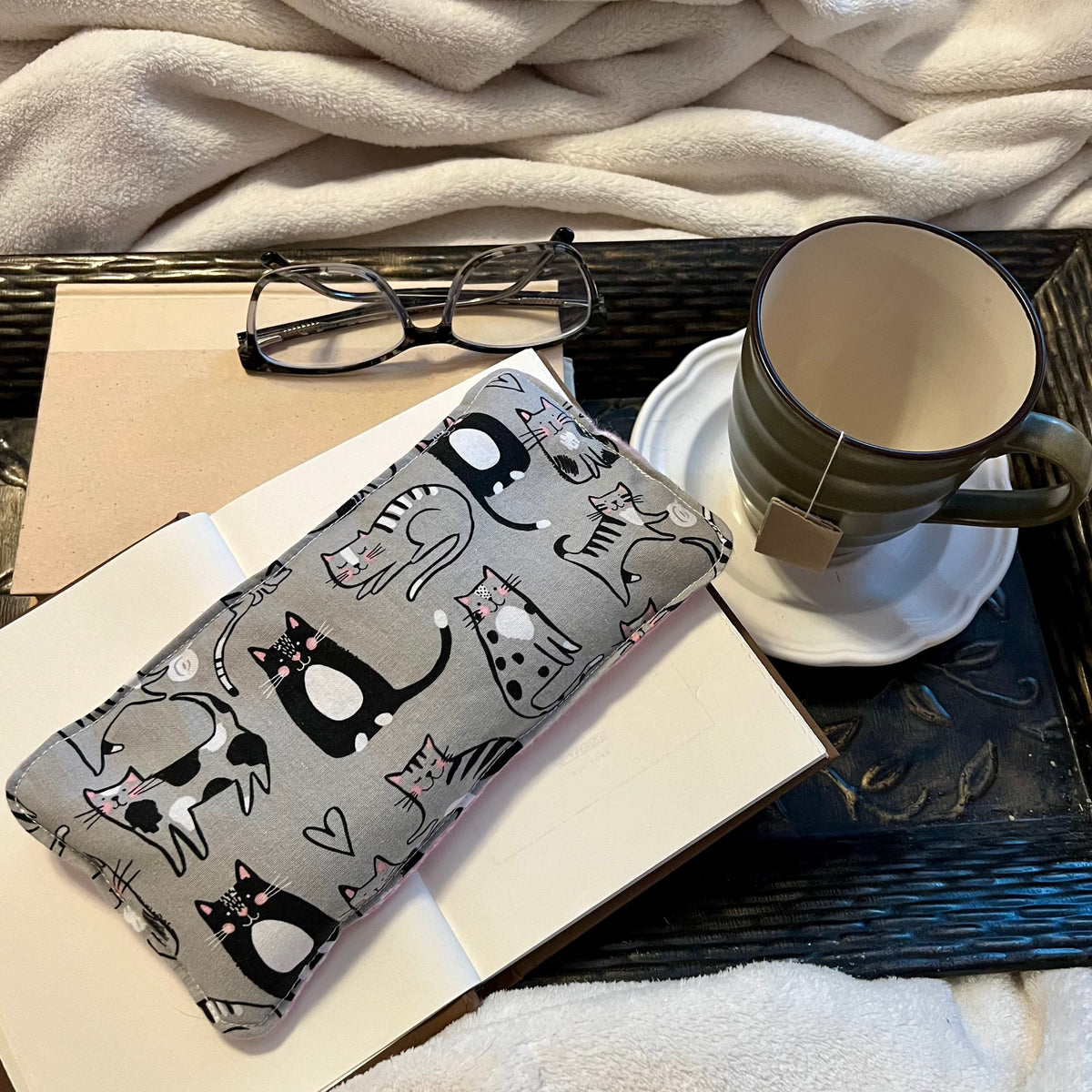 An Eclectic Spruce Purrfect Pause Weighted Eye Pillow sitting on a tray with a book, mug, pair of glasses, and a cozy white blanket.