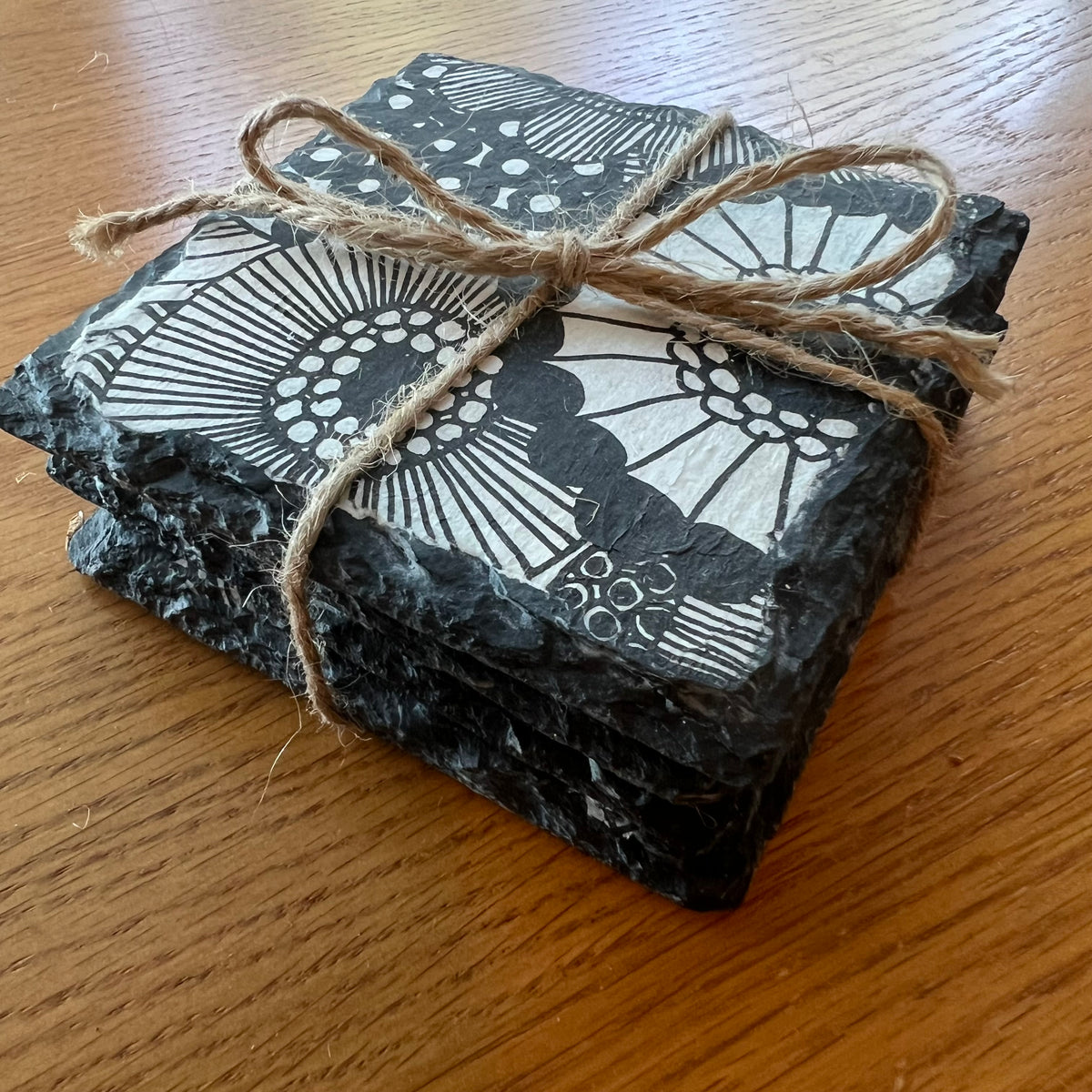 Four Eclectic Spruce black slate coasters tied with a rustic jute twine bow and set on a wooden tabletop. Each coaster is a 4x4 inch square and features a unique black and white doodle-style floral and circular design that is decoupaged onto the slate using an 8-step artisan process and industrial strength protective varnish.