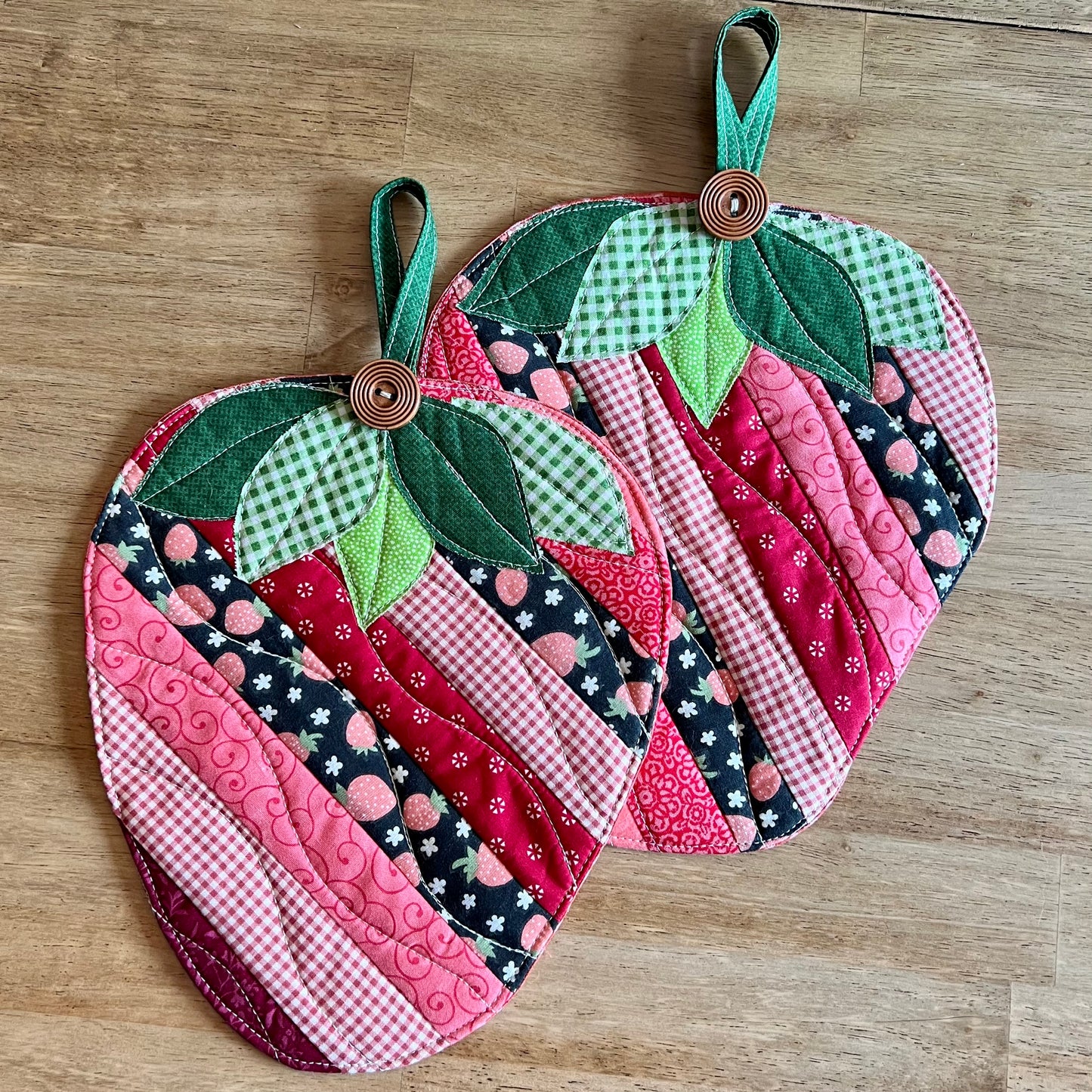 Two Eclectic Spruce Sweet Strawberry hot pads (strawberry shaped, quilted potholders with green leaf tops and wooden button handles), displayed on a wooden tabletop.