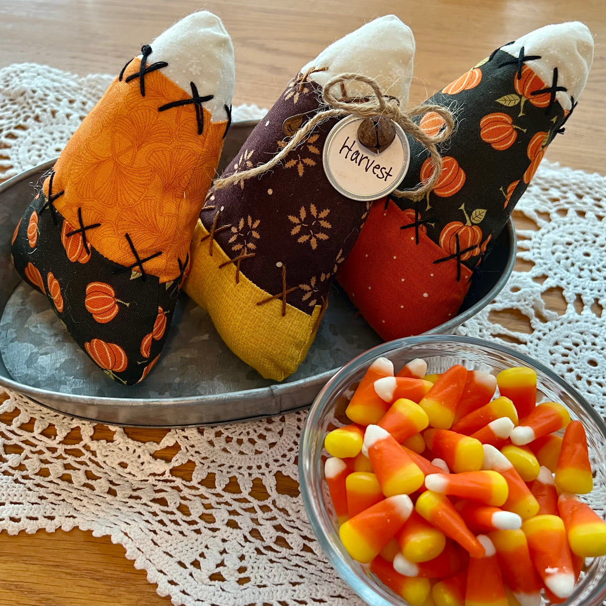 Three Eclectic Spruce handmade fabric candy corn bowl fillers with autumn harvest themes are arranged on a galvanized metal tray. The tray is sitting on a doily next to a glass bowl filled with candy corns.