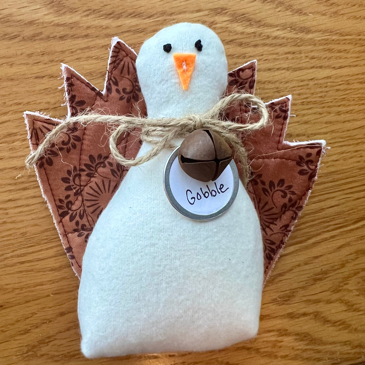 An Eclectic Spruce handmade fabric Thanksgiving turkey sitting on a wooden table. The turkey features a white flannel body, textile tail feathers, an orange felt beak, black stitched eyes, a brown rusty bell on a twine bow, and a round white tag that reads "Gobble."