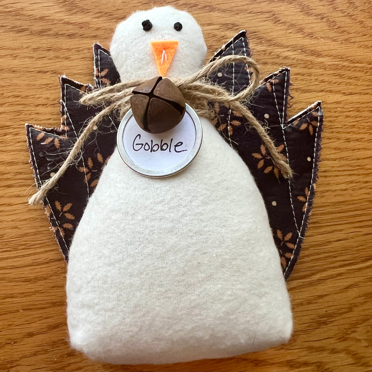 An Eclectic Spruce handmade fabric Thanksgiving turkey sitting on a wooden table. The turkey features a white flannel body, textile tail feathers, an orange felt beak, black stitched eyes, a brown rusty bell on a twine bow, and a round white tag that reads "Gobble."