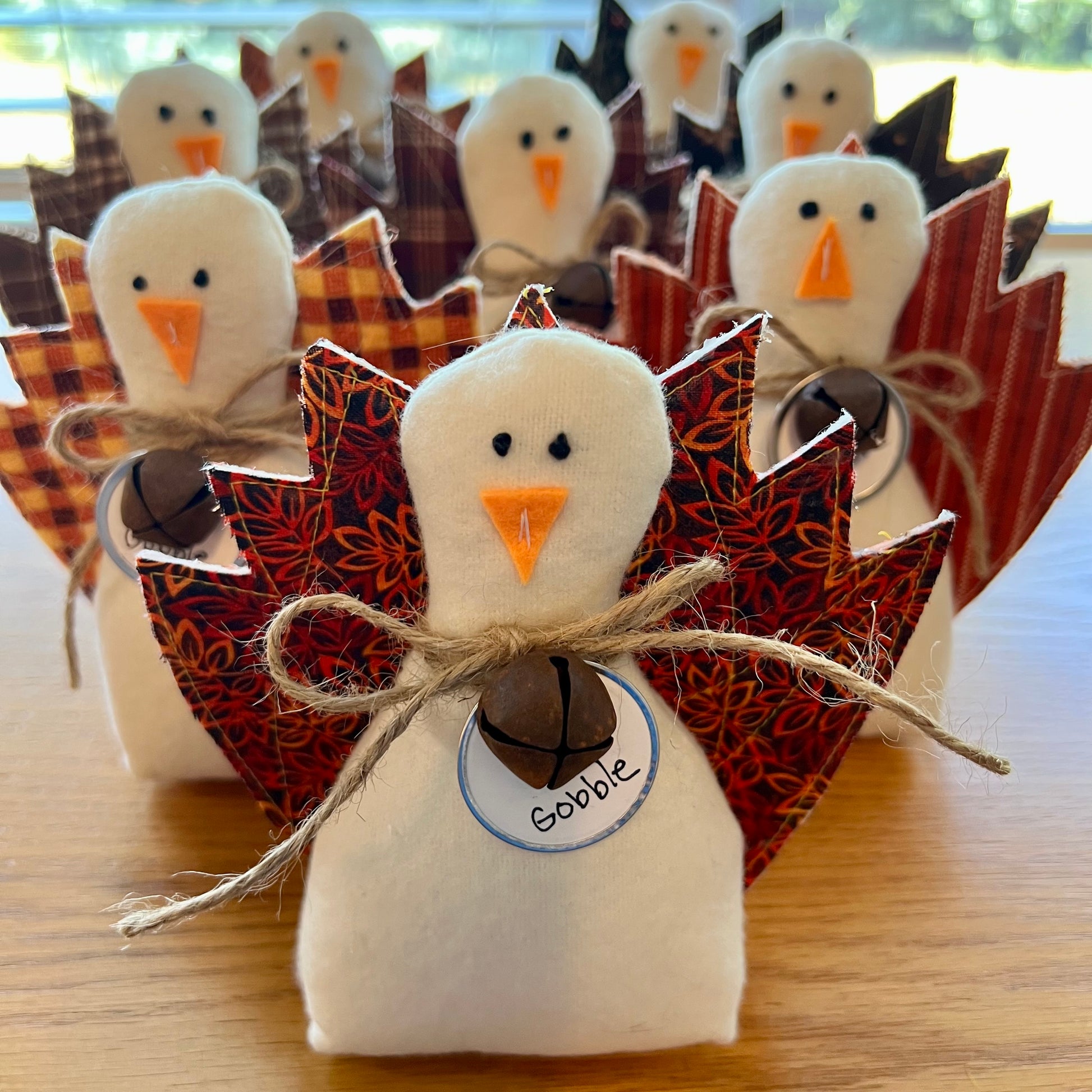 A set of Eclectic Spruce handmade fabric Thanksgiving turkeys arranged standing up on a wooden table. The center turkey is in sharp focus, made with a white flannel body, textile tail feathers, an orange felt beak, black stitched eyes, a brown rusty bell on a twine bow, and a round white tag that reads "Gobble."