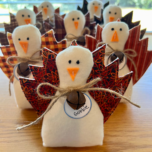 A set of Eclectic Spruce handmade fabric Thanksgiving turkeys arranged standing up on a wooden table. The center turkey is in sharp focus, made with a white flannel body, textile tail feathers, an orange felt beak, black stitched eyes, a brown rusty bell on a twine bow, and a round white tag that reads "Gobble."