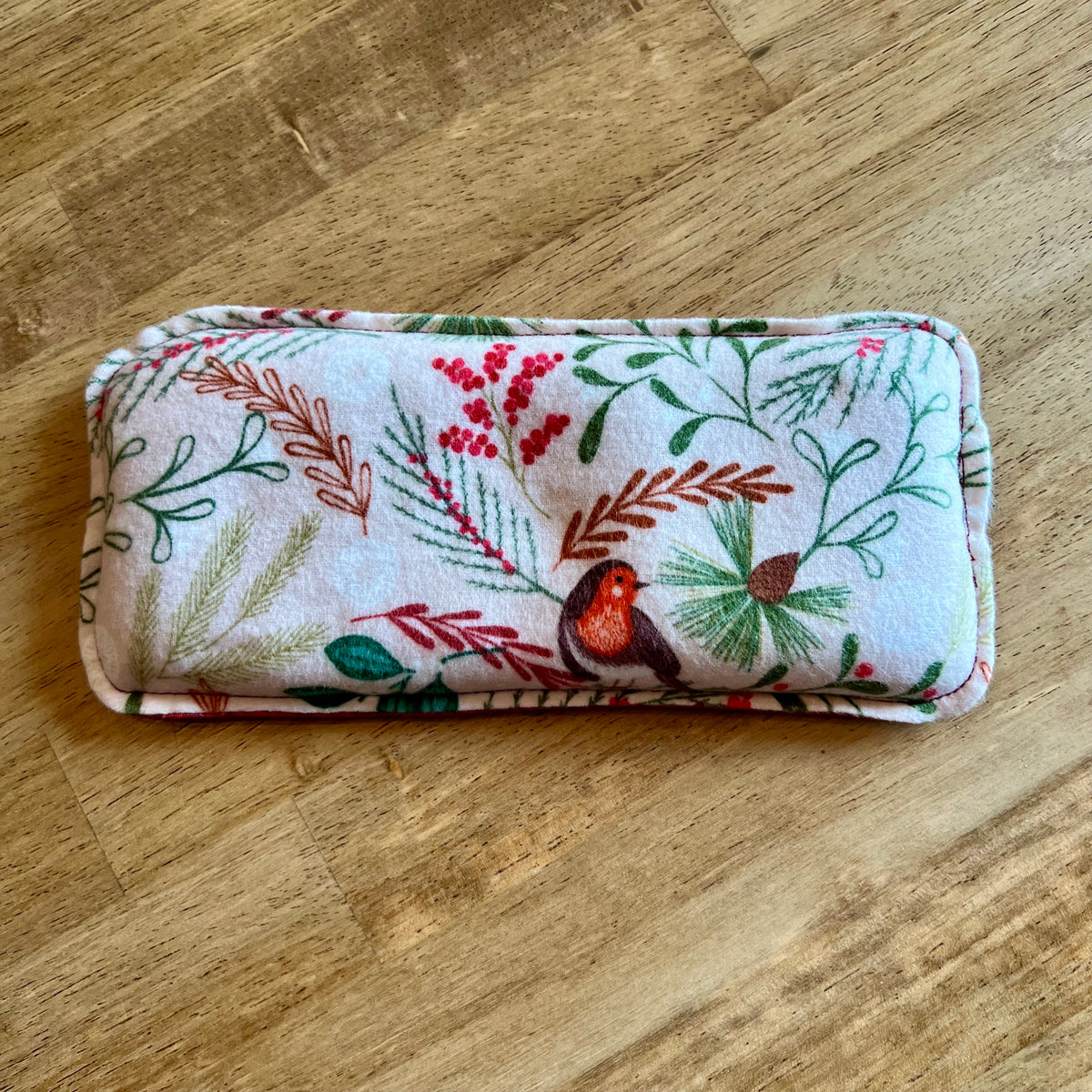 A dual-sided Woodland Bough Weighted Eye Pillow by Eclectic Spruce. This pillow features a winter-themed print of a robin bird, red berries, and green foliage on a cream background.