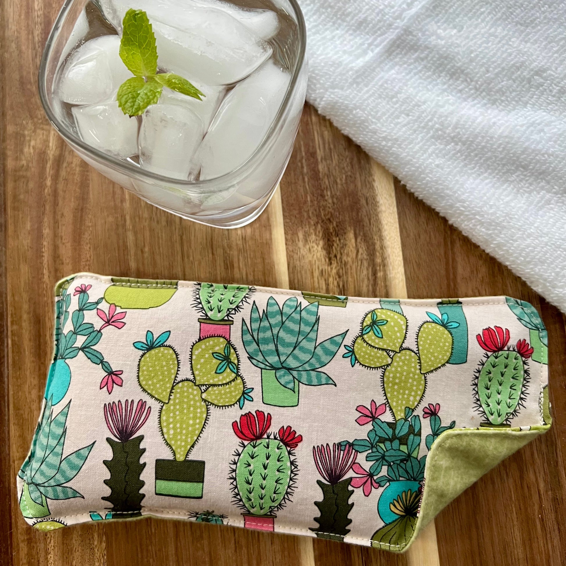 Weighted Eye Pillow on a wood surface next to a white towel and glass of ice water.