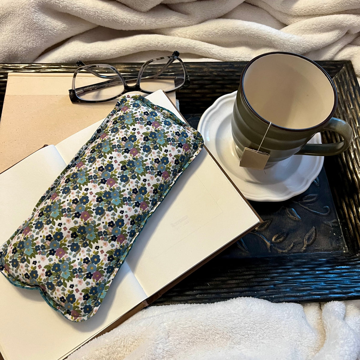 An Eclectic Spruce Wildflower Glen Weighted Eye Pillow sitting on a tray with a book, mug, pair of glasses, and a cozy white blanket.