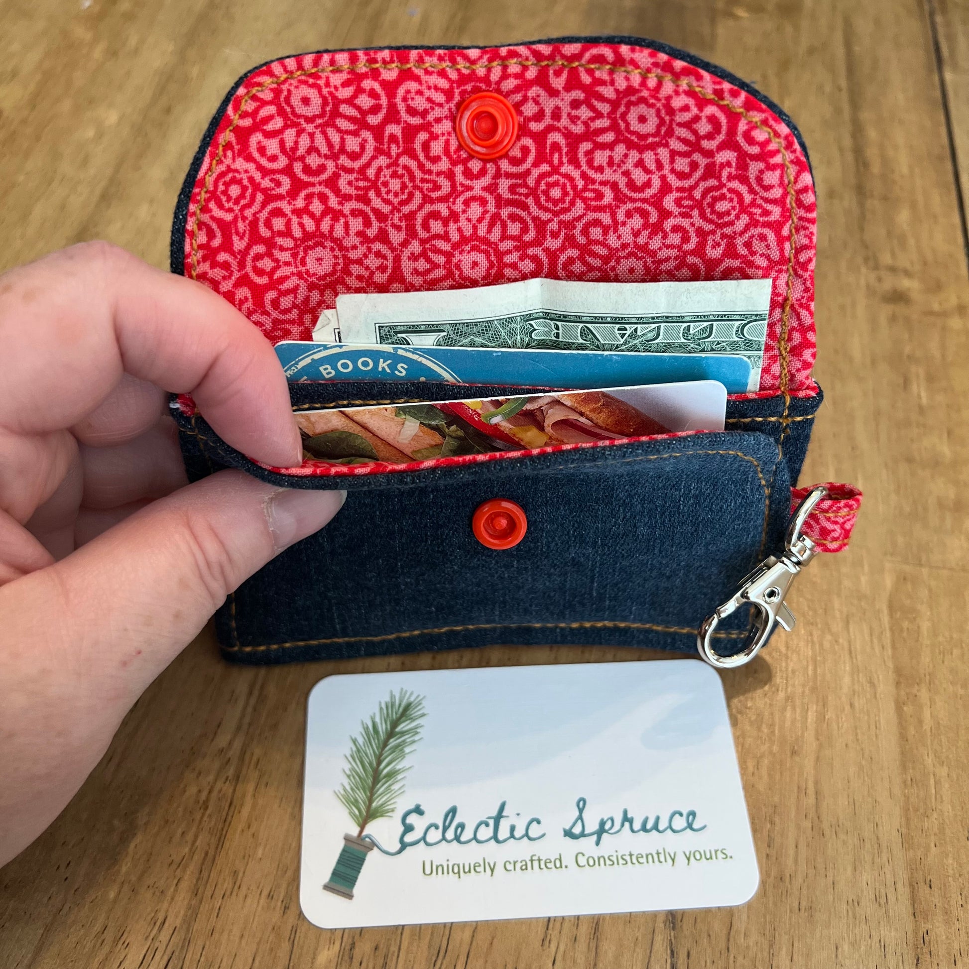 Eclectic Spruce handcrafted mini card holder wallet with a dark blue denim exterior and a red floral patterned interior, shown open with cash and cards inside two card slots. The wallet contains a bright red snap closure and a silver metal swivel clasp for convenience and security.