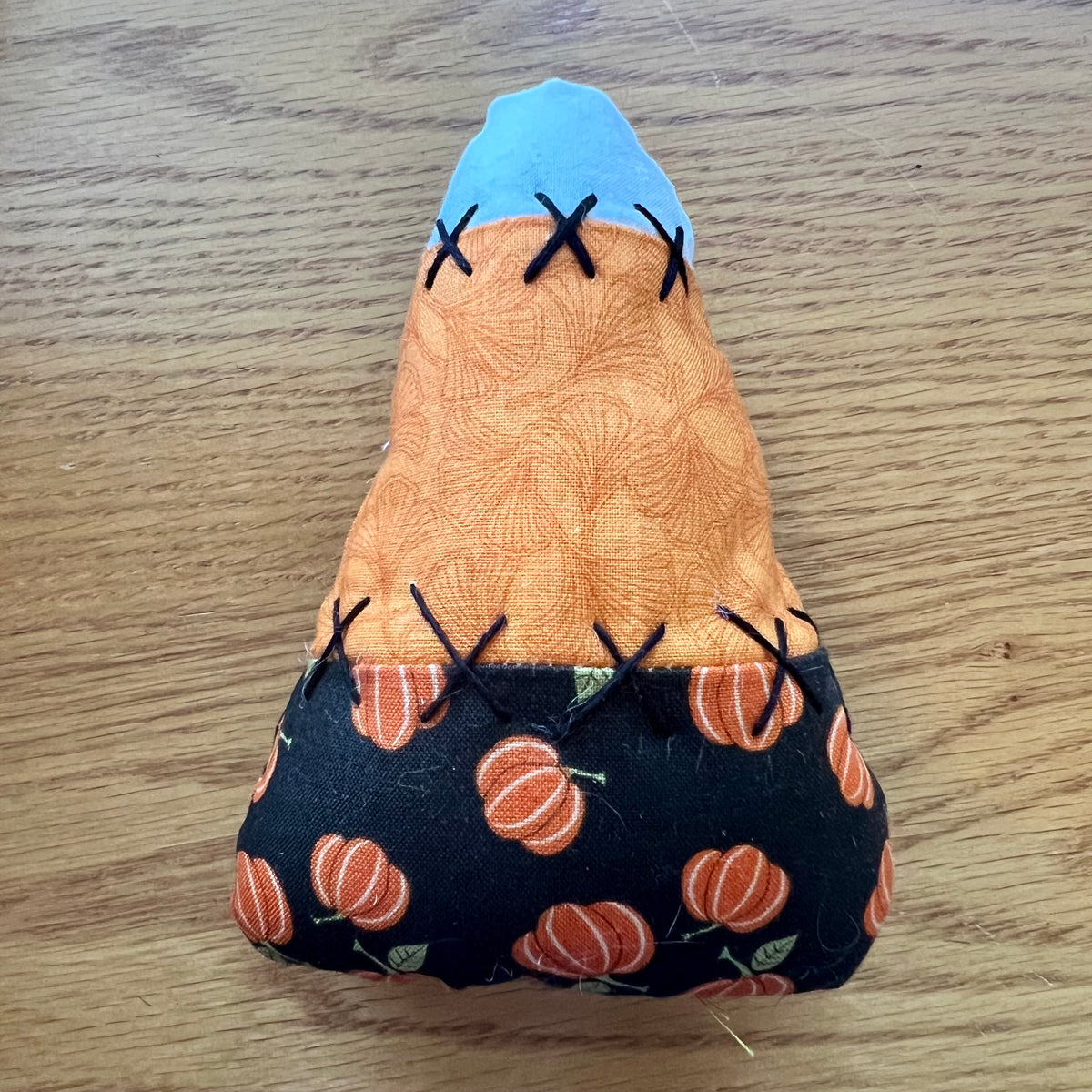 An Eclectic Spruce handmade fabric candy corn, featuring black fabric with a pumpkin pattern on the bottom, a bright orange middle section, and a white top section. It is decorated with hand stitched details.