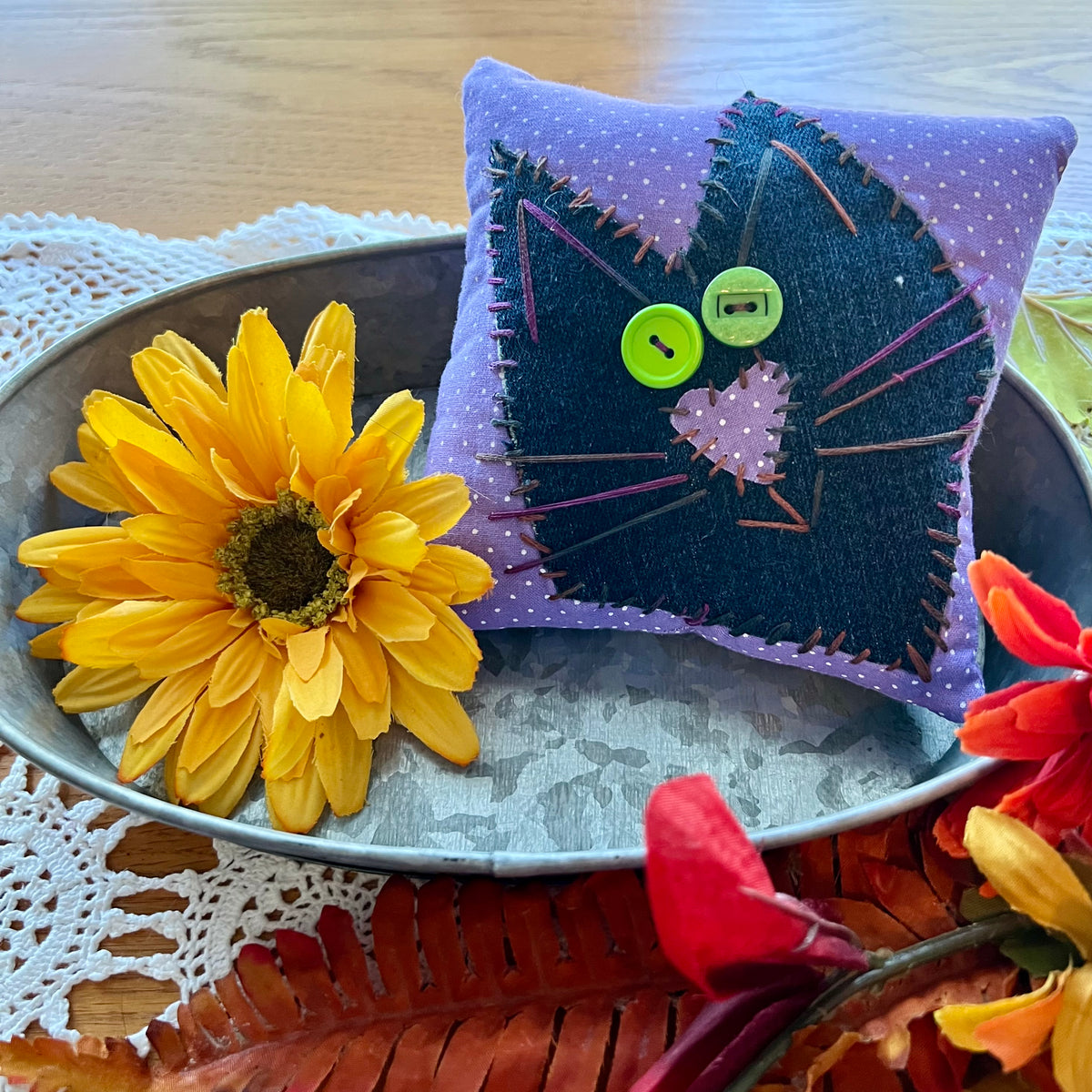 One handmade Eclectic Spruce Purrfect Patchwork Cat pillows styled in an autumn setting. The pillow features a black cat face applique on an purple fabric with white polka dots, complete with green mismatched button eyes, a stitched applique nose, and stitched whiskers.