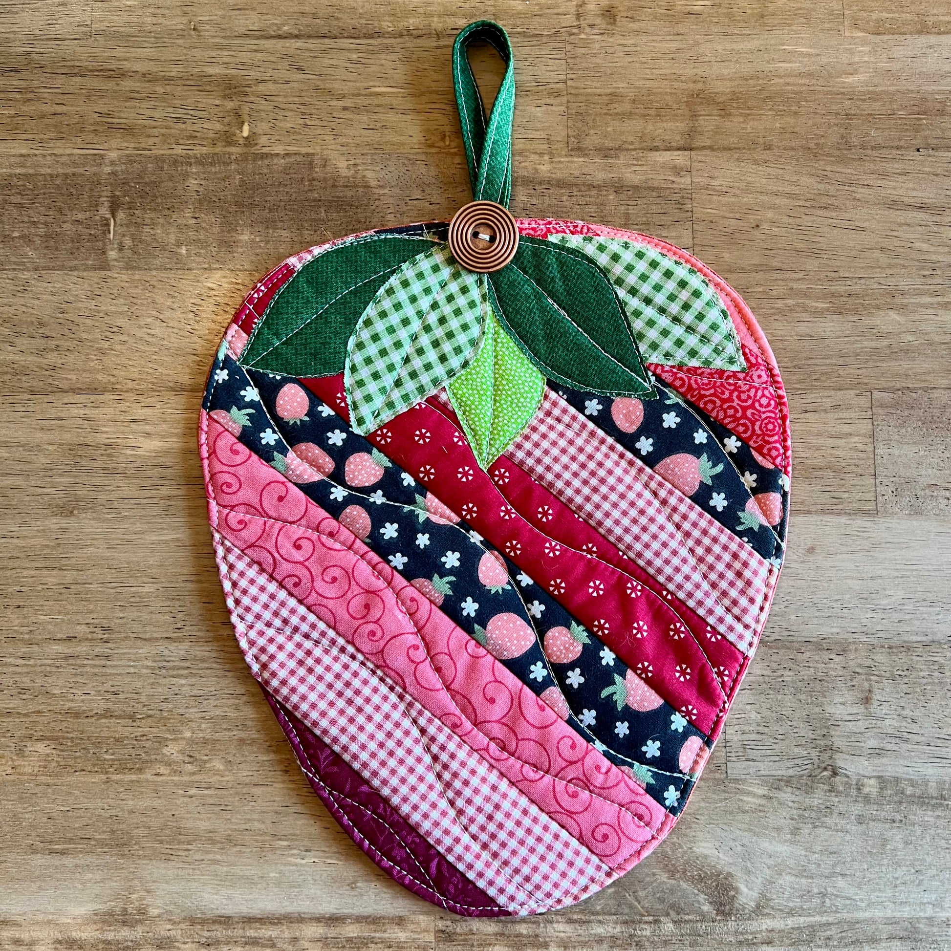 One strawberry-shaped quilted potholder on a wooden table. This is the   Eclectic Spruce Sweet Strawberry hot pad, which features strip patchwork in various pink, red, white, and coral cotton prints, green leaves, and a wooden button accent.