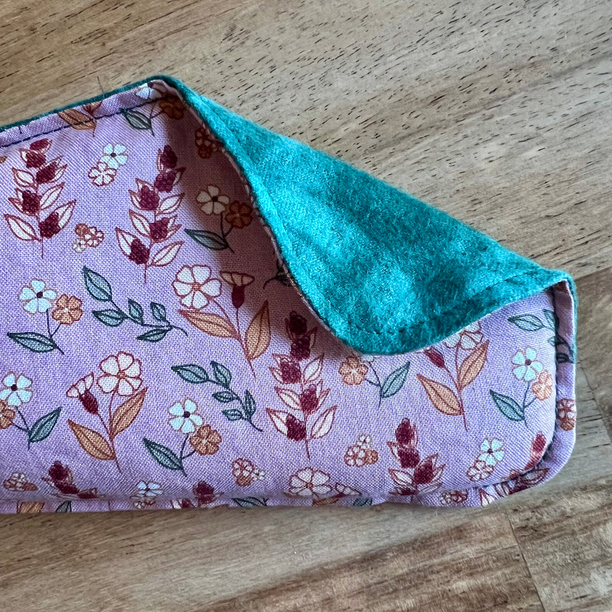 A close-up of the Evening Primrose Weighted Eye Pillow by Eclectic Spruce. The front side is a mauve cotton fabric with a floral pattern in red, green, white, and gold. A corner of the pillow is folded over, revealing a textured teal green flannel fabric on the reverse side.