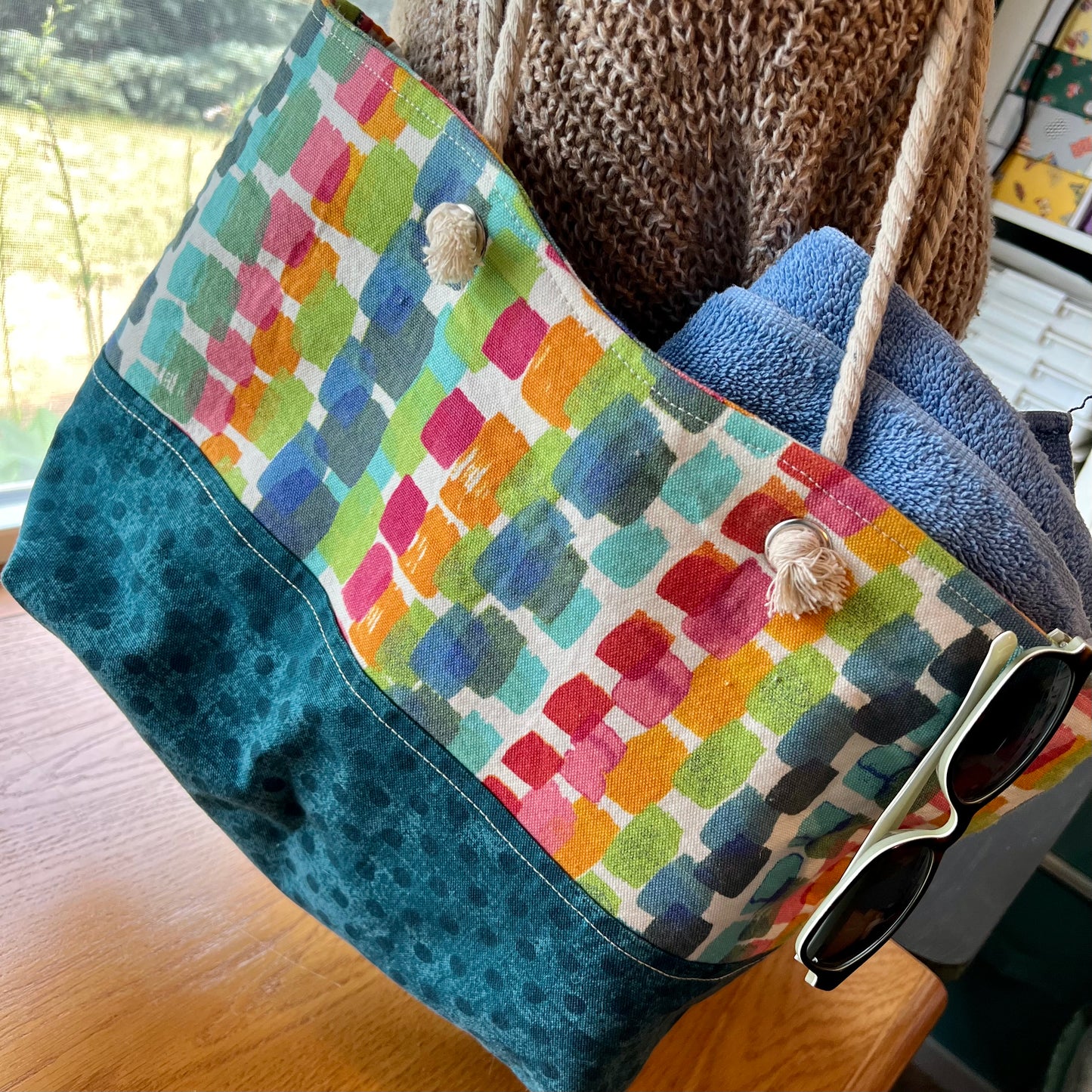 Eclectic Spruce handmade rope-handled tote bag - a large tote bag with a colorful geometric pattern on the upper half and a teal polka dot lower half, shown with a blue towel and sunglasses inside.