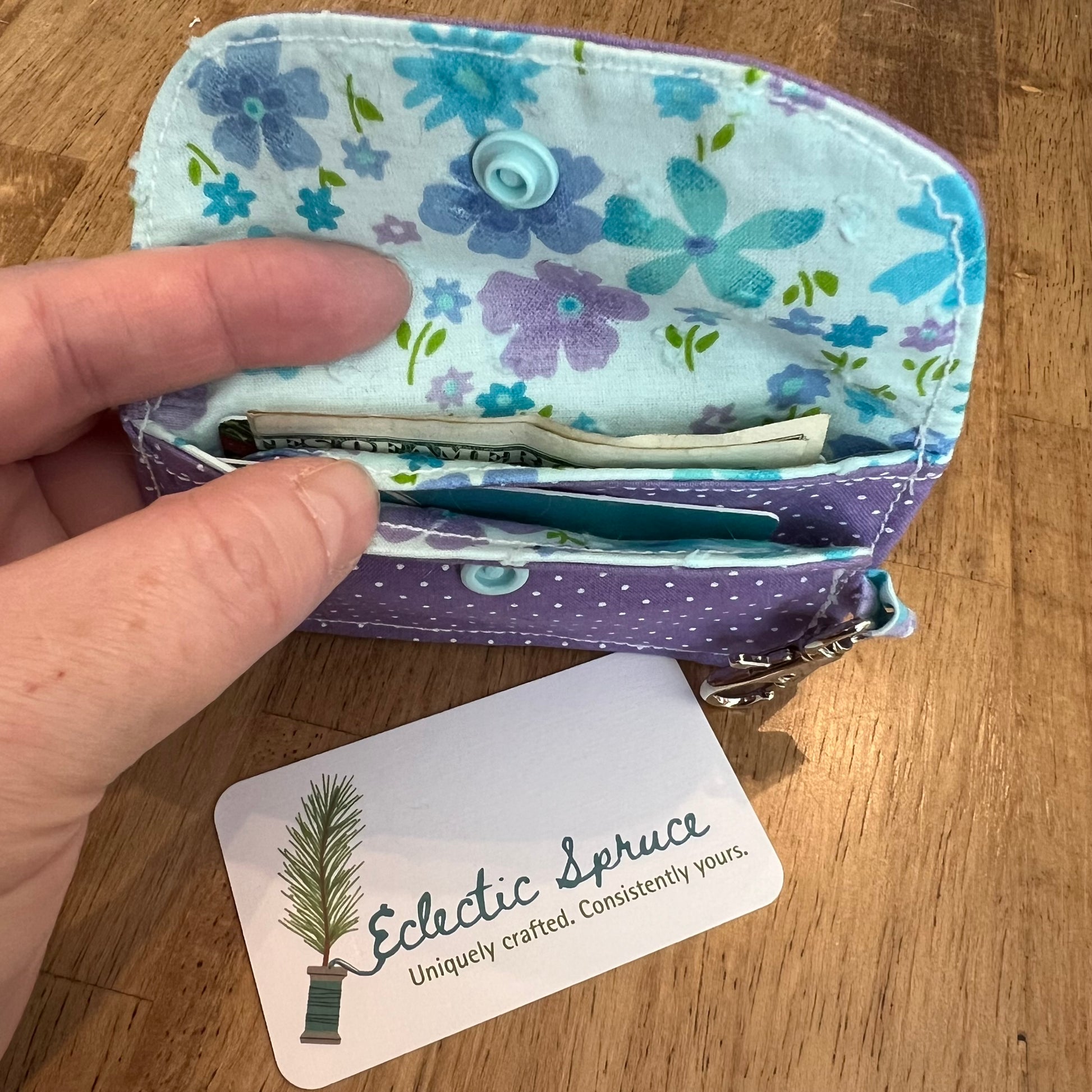 Eclectic Spruce handcrafted mini card holder wallet with a purple polka dot patterned exterior and a light blue floral patterned interior, shown open with cash and cards inside two card slots. The wallet contains a sky blue snap closure and a silver metal swivel clasp for convenience and security.
