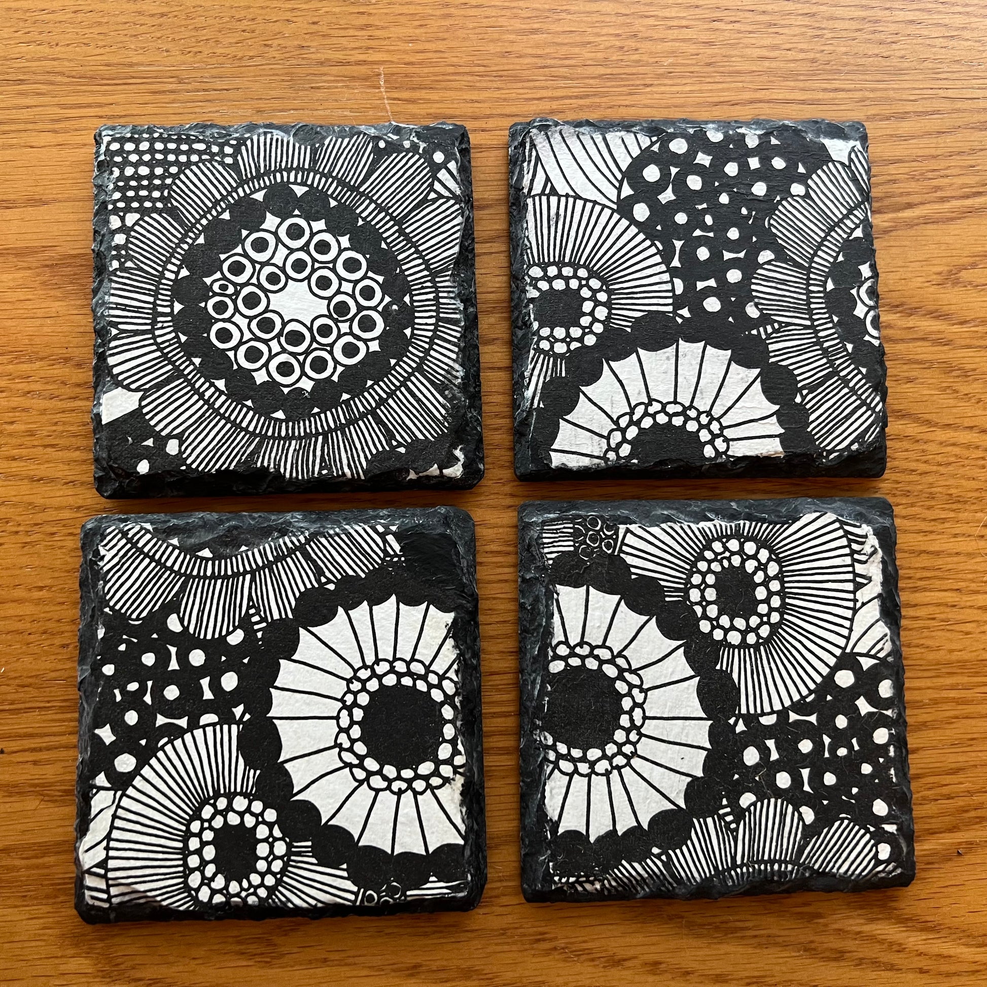 Four Eclectic Spruce slate coasters arranged in a 2x2 grid on a wooden tabletop. Each coaster is a 4x4 inch square and features a unique black and white doodle-style floral and circular design that is decoupaged onto the slate using an 8-step artisan process and industrial strength protective varnish.