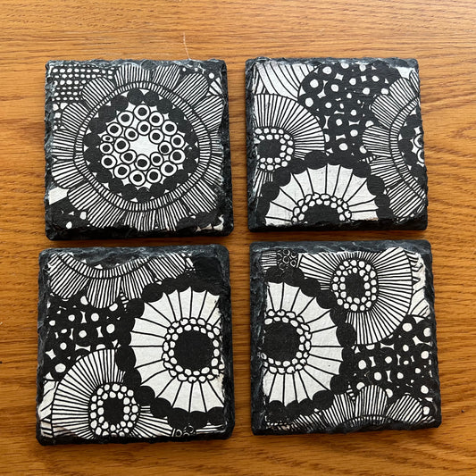 Four Eclectic Spruce slate coasters arranged in a 2x2 grid on a wooden tabletop. Each coaster is a 4x4 inch square and features a unique black and white doodle-style floral and circular design that is decoupaged onto the slate using an 8-step artisan process and industrial strength protective varnish.
