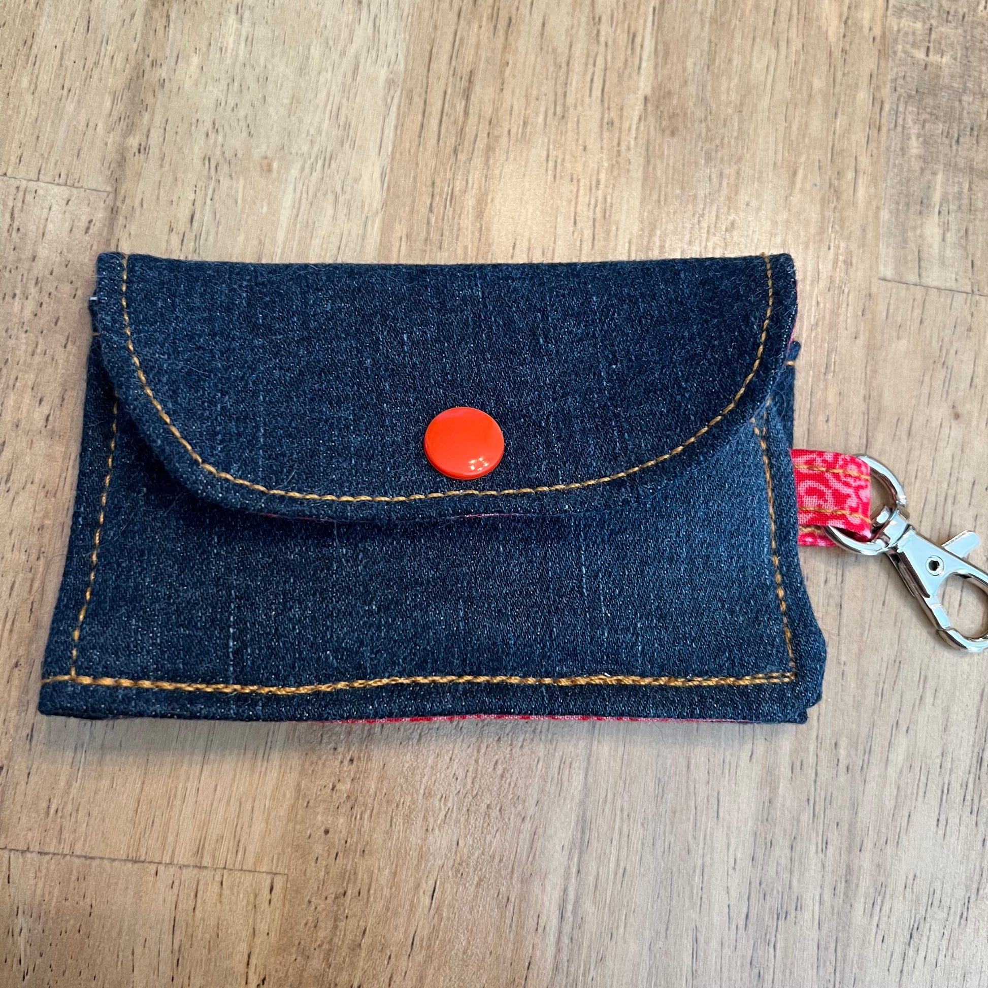 Eclectic Spruce handcrafted mini snap wallet keychain with snap button closure made of dark upcycled denim with orange contrast stitching, a bright red snap button, red floral fabric loop, a silver metal swivel clasp. 