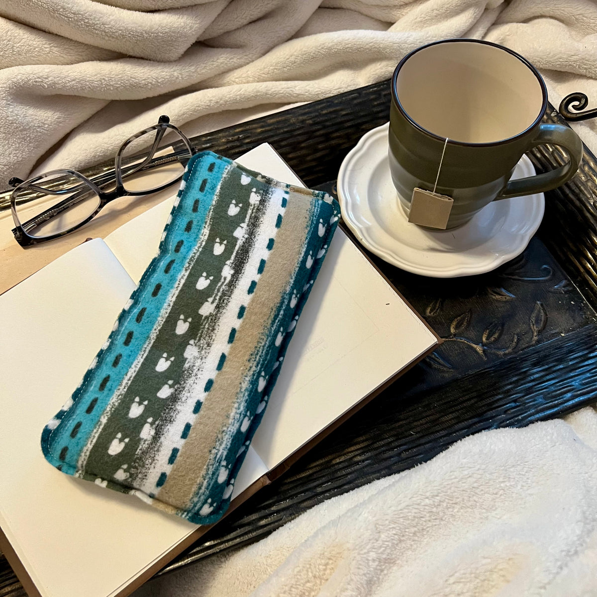 An Eclectic Spruce Alpine Path Weighted Eye Pillow sitting on a tray with a book, mug, pair of glasses, and a cozy white blanket.