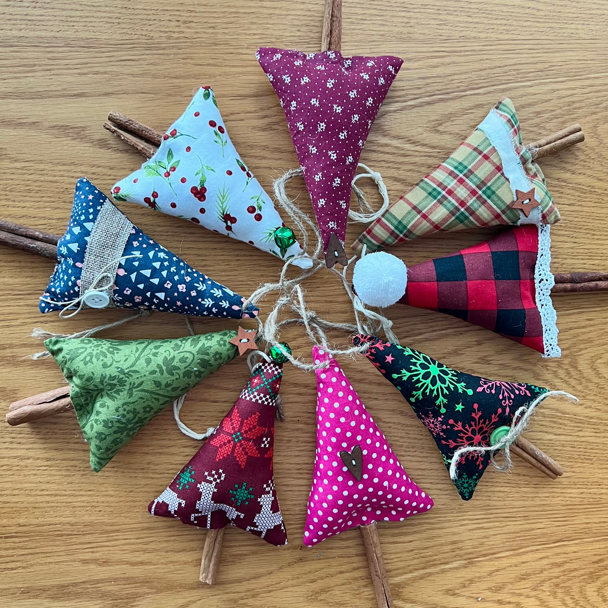 Eclectic Spruce's handmade cinnamon stick trees are made with cotton fabric, stuffed with fiberfill, and embellished with real cinnamon sticks along with lace, buttons, bells, and other trim.  Sold mystery style, you will receive a unique tree (or curated combination of trees) picked out just for  you. This photo shows 8 Eclectic Spruce trees arranged in a circle on a wood surface.