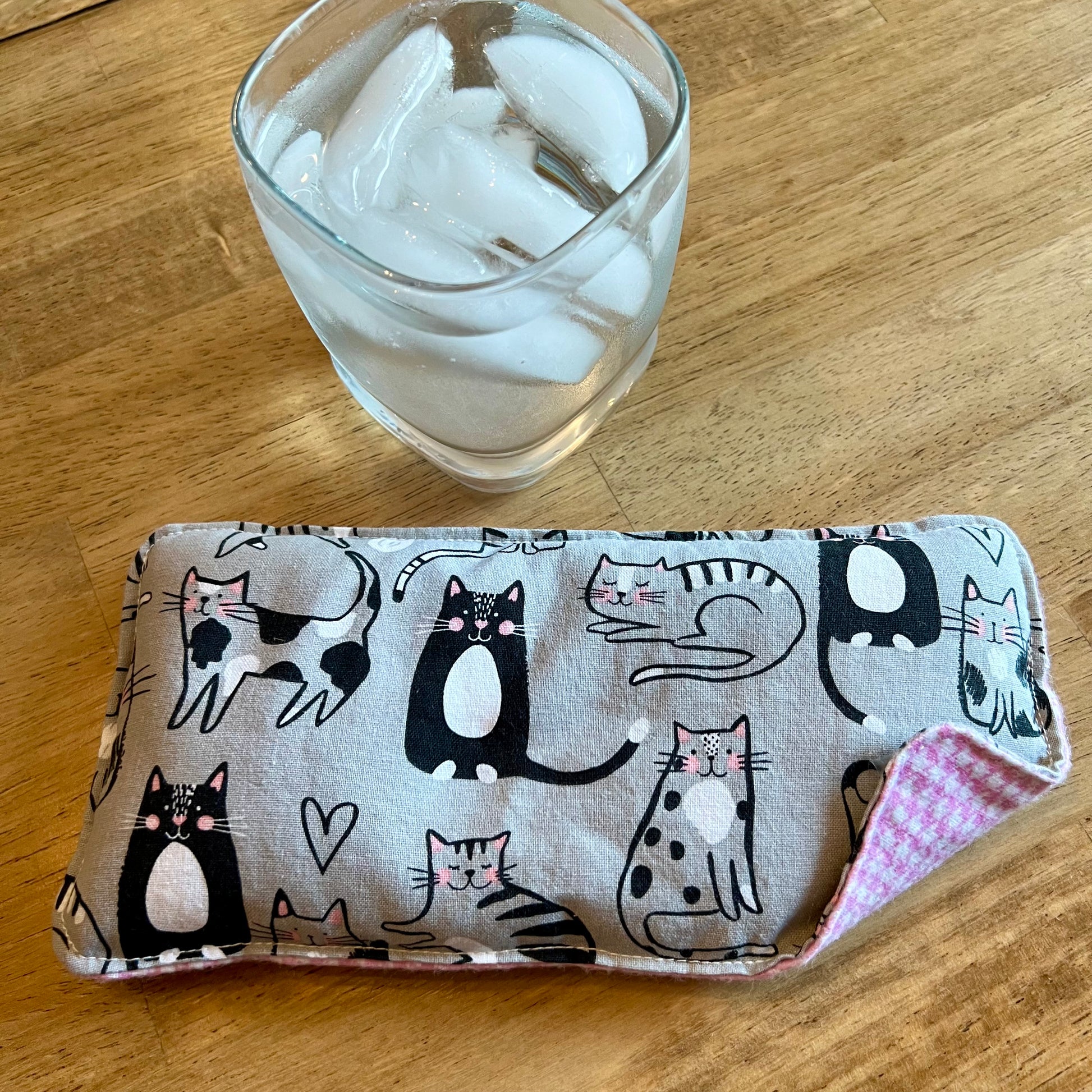 An Eclectic Spruce Purrfect Pause Weighted Eye pillow on a wood surface next to a glass of ice water.