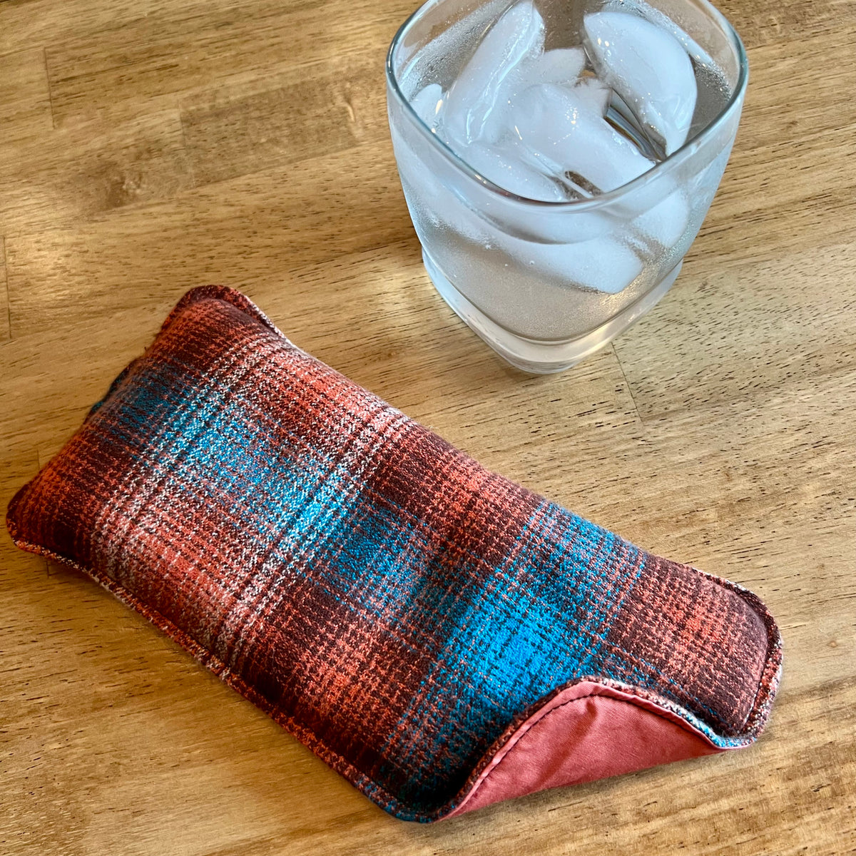 An Eclectic Spruce Copper Ridge Weighted Eye pillow on a wood surface next to a glass of ice water.