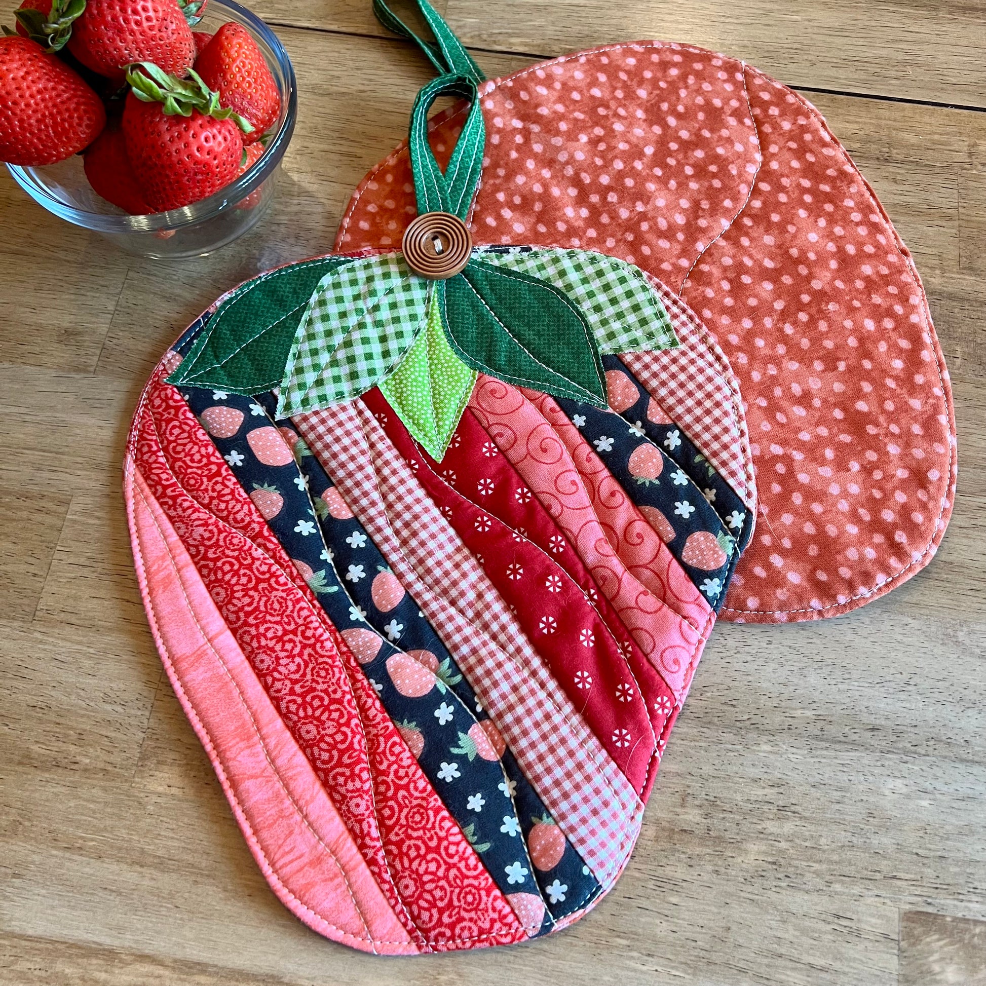 Two strawberry-shaped quilted potholders on a wooden table, with a small glass bowl of fresh strawberries in the background. One potholder is flipped upside down showing the "strawberry print" backside to the potholder. The other one features the front side of the Eclectic Spruce Sweet Strawberry hot pad, which features strip patchwork in various pink, red, white, and coral cotton prints, green leaves, and a wooden button accent.
