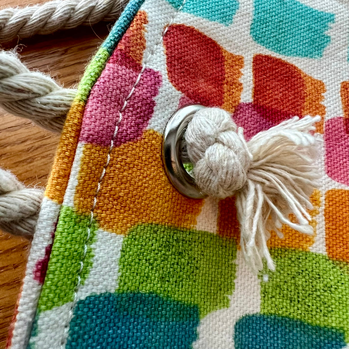 Close-up of the Eclectic Spruce rope-handled tote bag, with a colorful geometric pattern in orange, pink, green, teal, and white, featuring a white rope handle, metal grommet, and matching rope tassel.