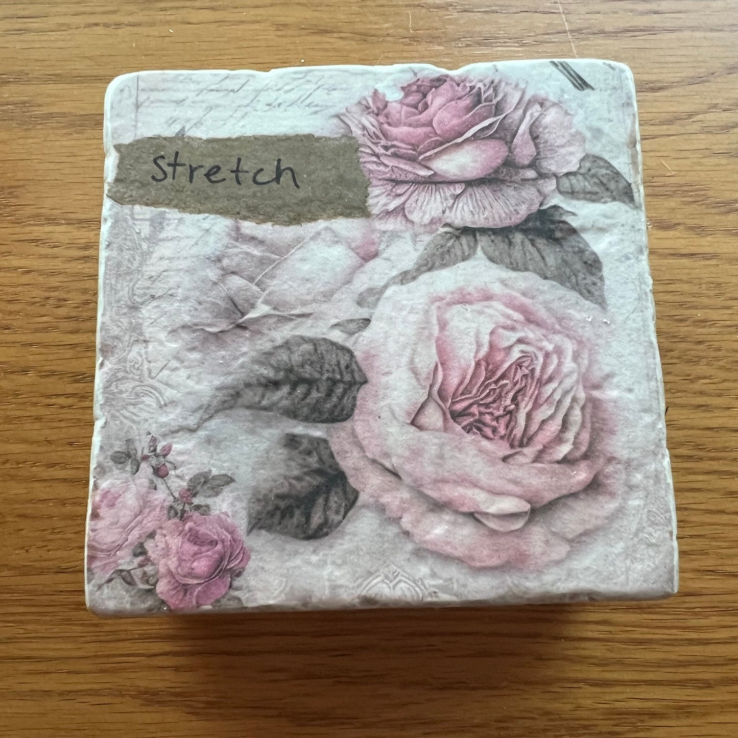 A top view of an Eclectic Spruce travertine coaster. It features a vintage floral design with pink roses and dark leaves, placed on a wooden surface. The top left corner has a brown tab with the word 'stretch' printed on it.