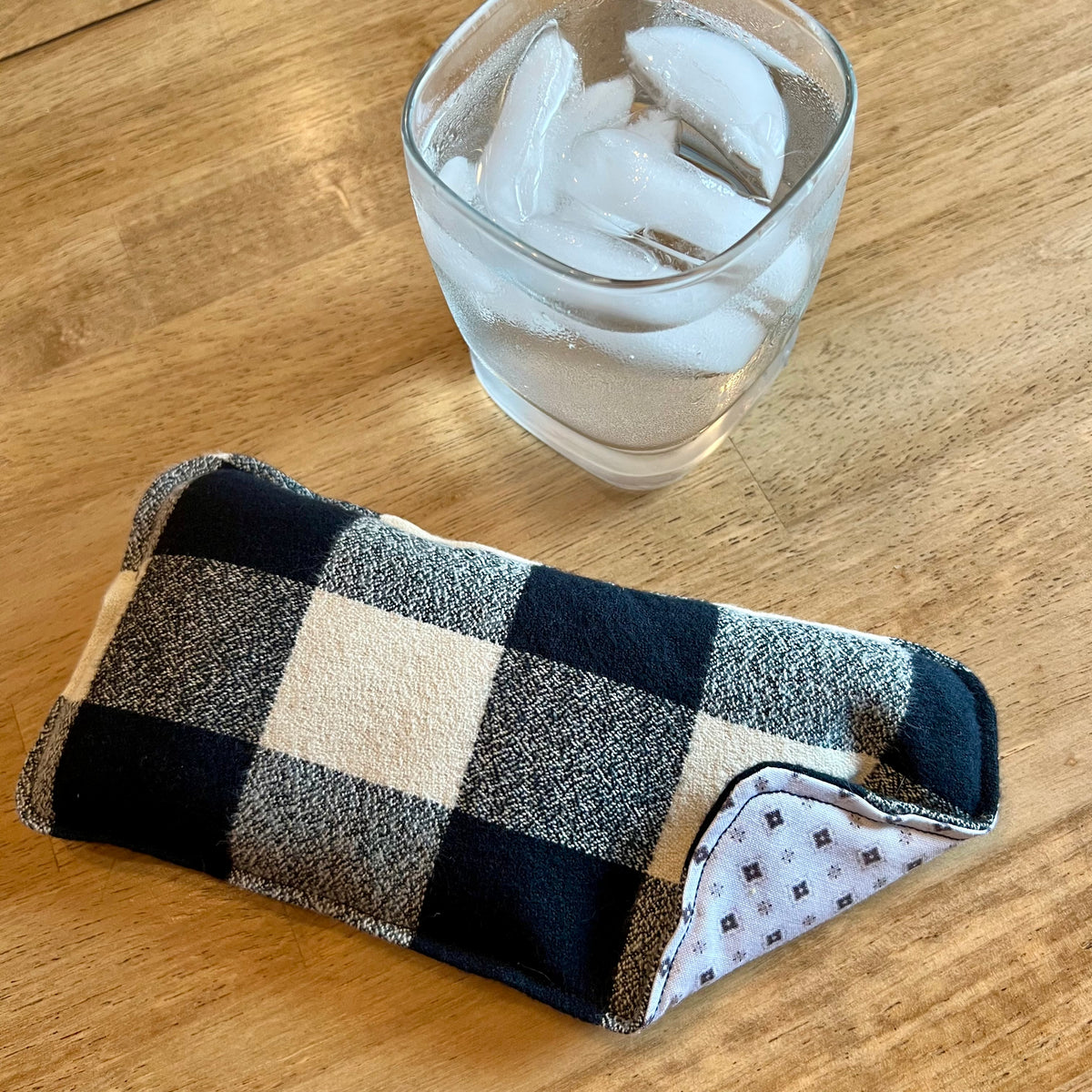 An Eclectic Spruce Nordic Check Weighted Eye pillow on a wood surface next to a glass of ice water.