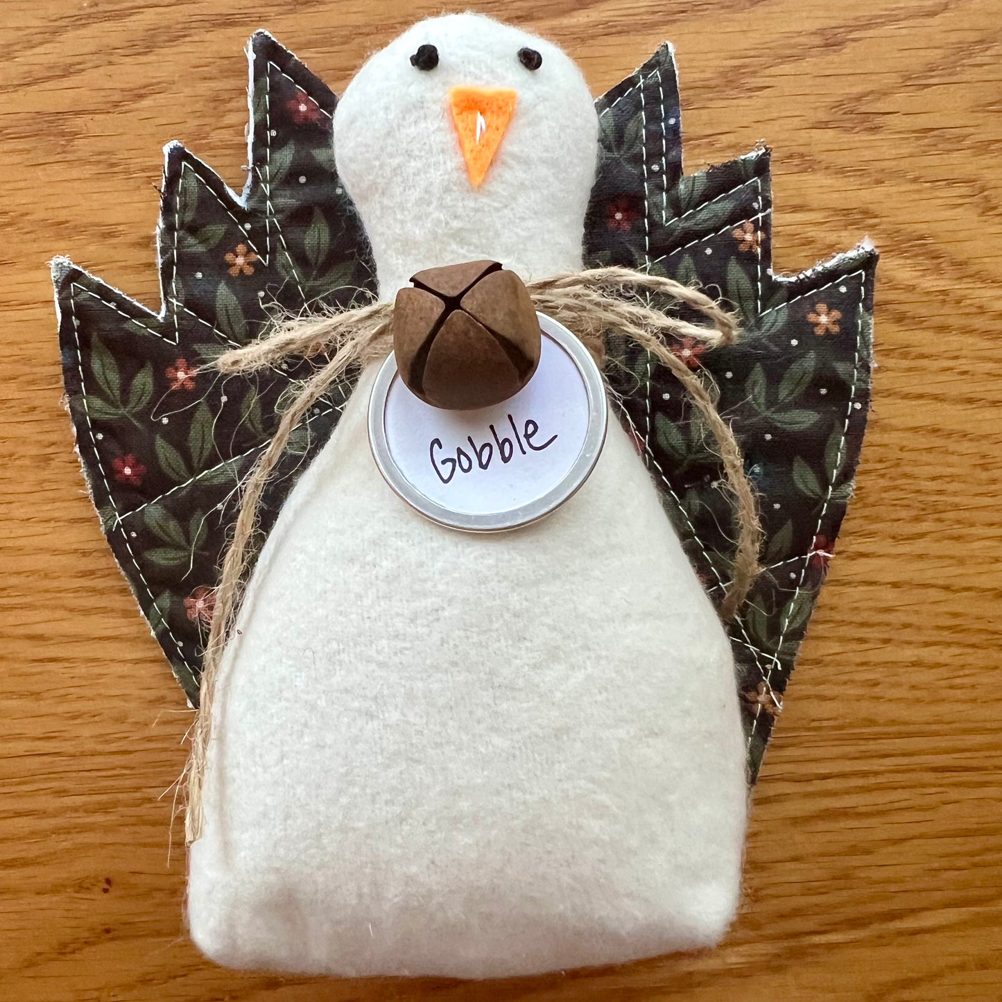 An Eclectic Spruce handmade fabric Thanksgiving turkey sitting on a wooden table. The turkey features a white flannel body, textile tail feathers, an orange felt beak, black stitched eyes, a brown rusty bell on a twine bow, and a round white tag that reads "Gobble."