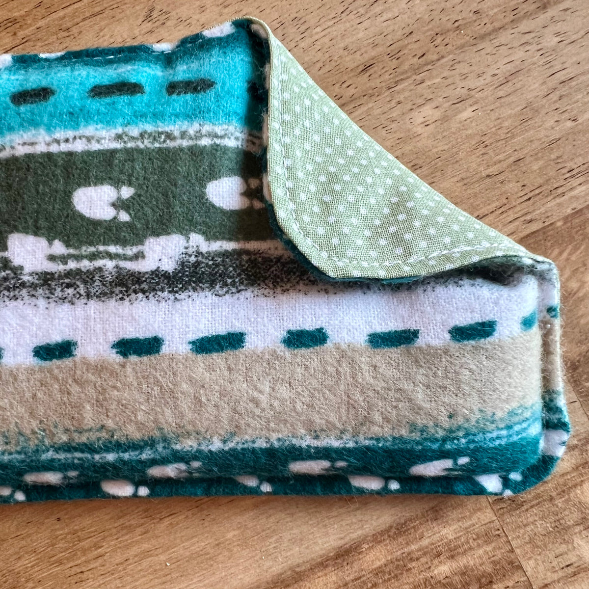 A close-up of the Alpine Path Weighted Eye Pillow by Eclectic Spruce. The front side features a flannel print containing teal, green, beige, and white stripes with animal tracks. One corner of the pillow if folded over, revealing a light green polka-dot cotton fabric on the reverse side.