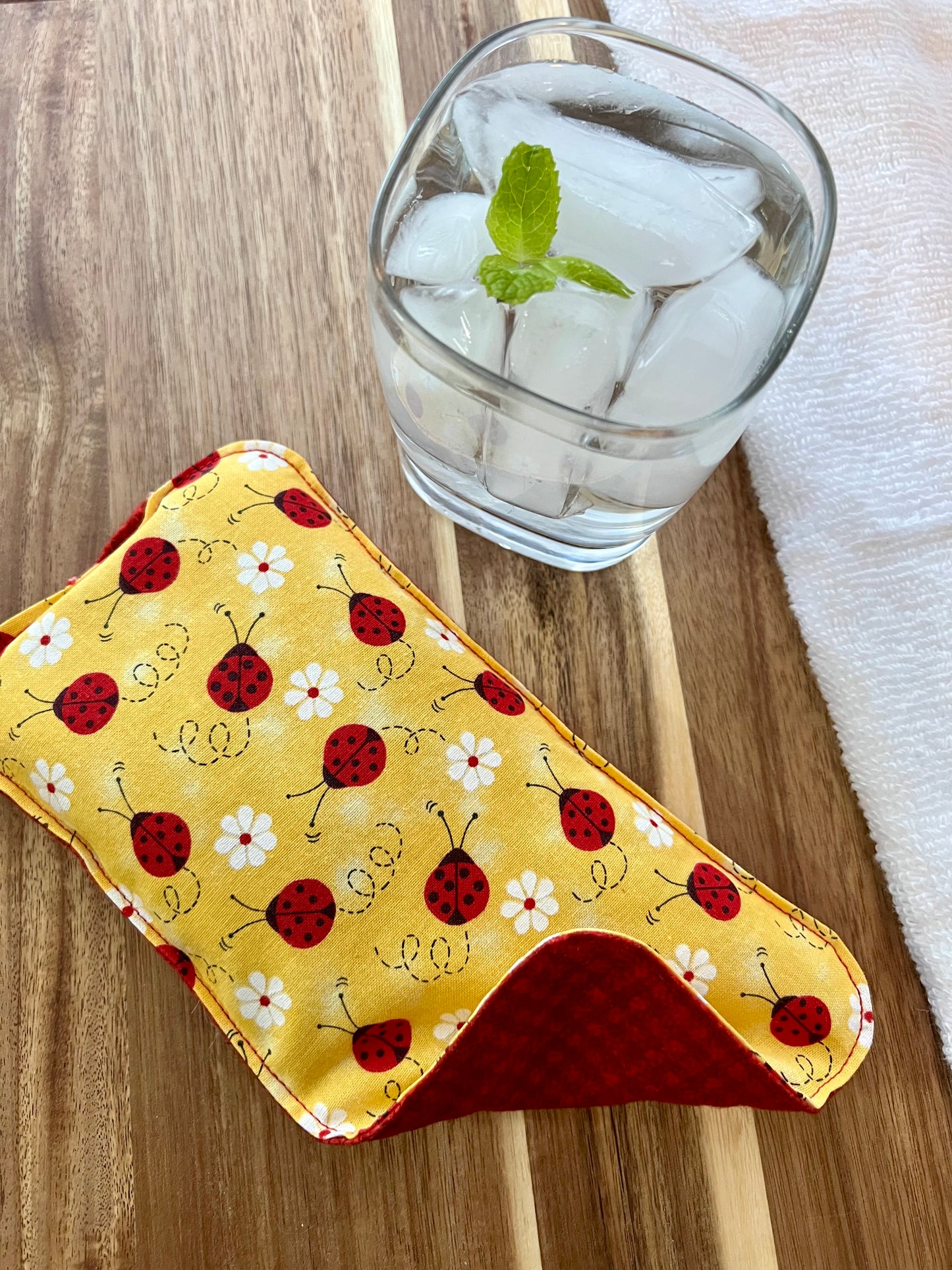 Weighted Eye Pillow on a wood surface next to a white towel and glass of ice water.