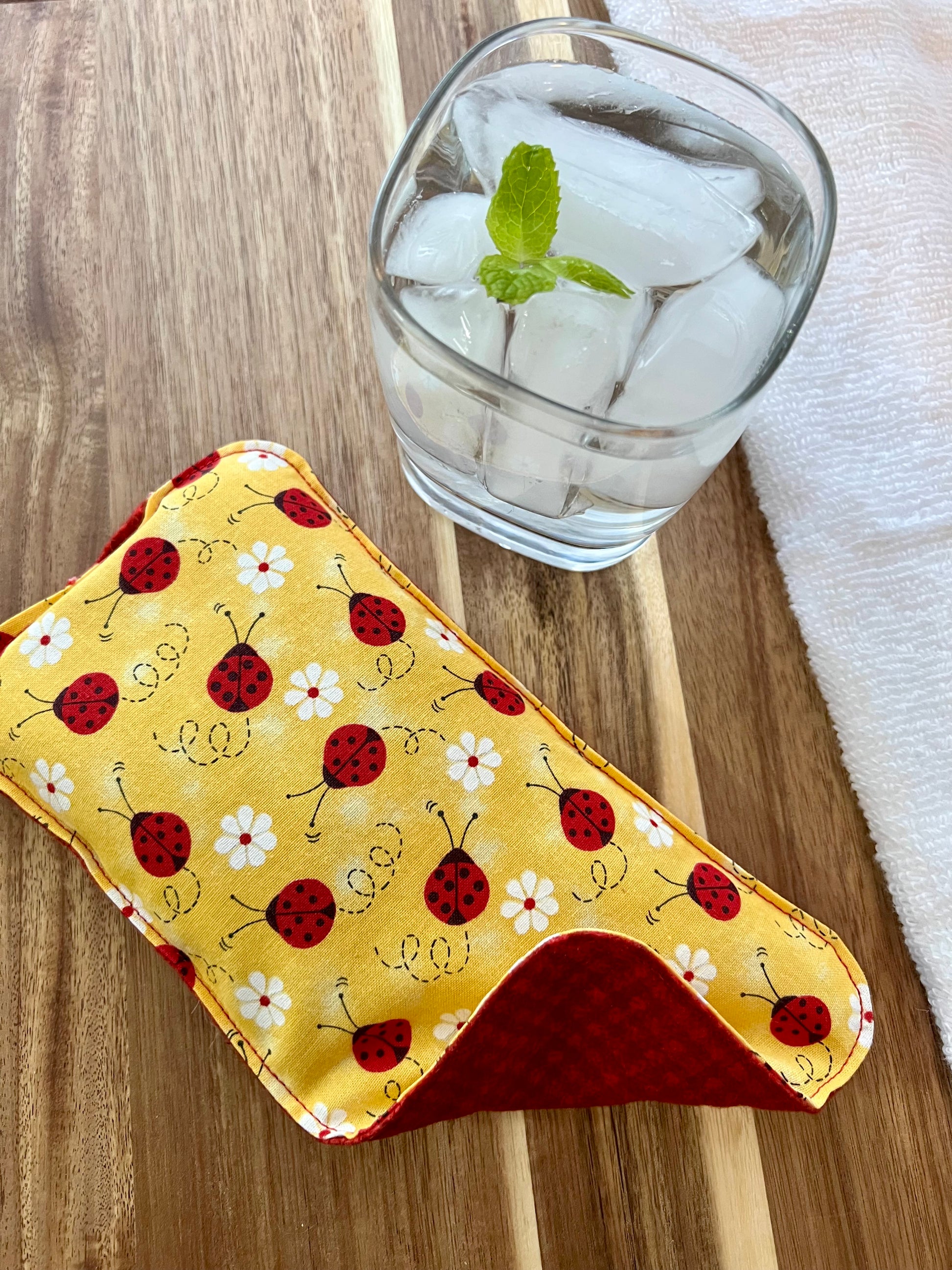 Weighted Eye Pillow on a wood surface next to a white towel and glass of ice water.