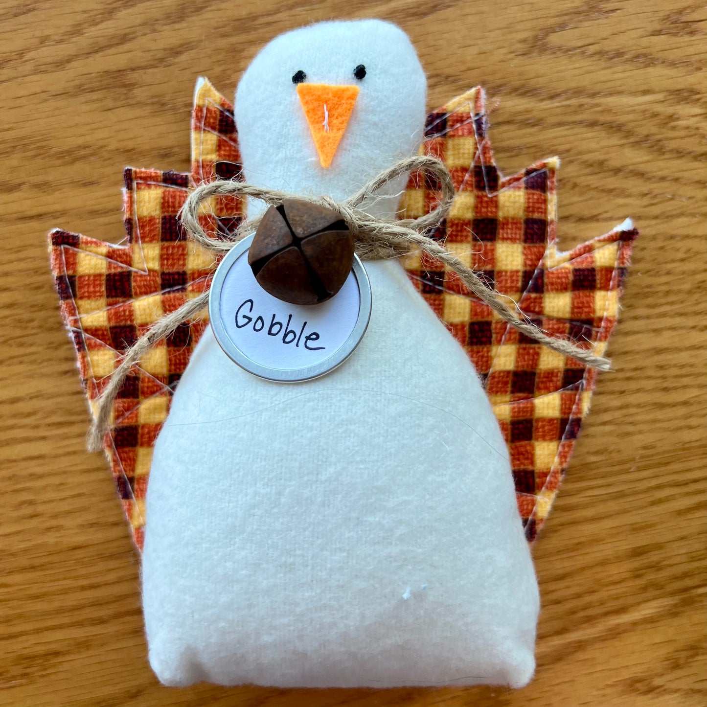 An Eclectic Spruce handmade fabric Thanksgiving turkey sitting on a wooden table. The turkey features a white flannel body, textile tail feathers, an orange felt beak, black stitched eyes, a brown rusty bell on a twine bow, and a round white tag that reads "Gobble."