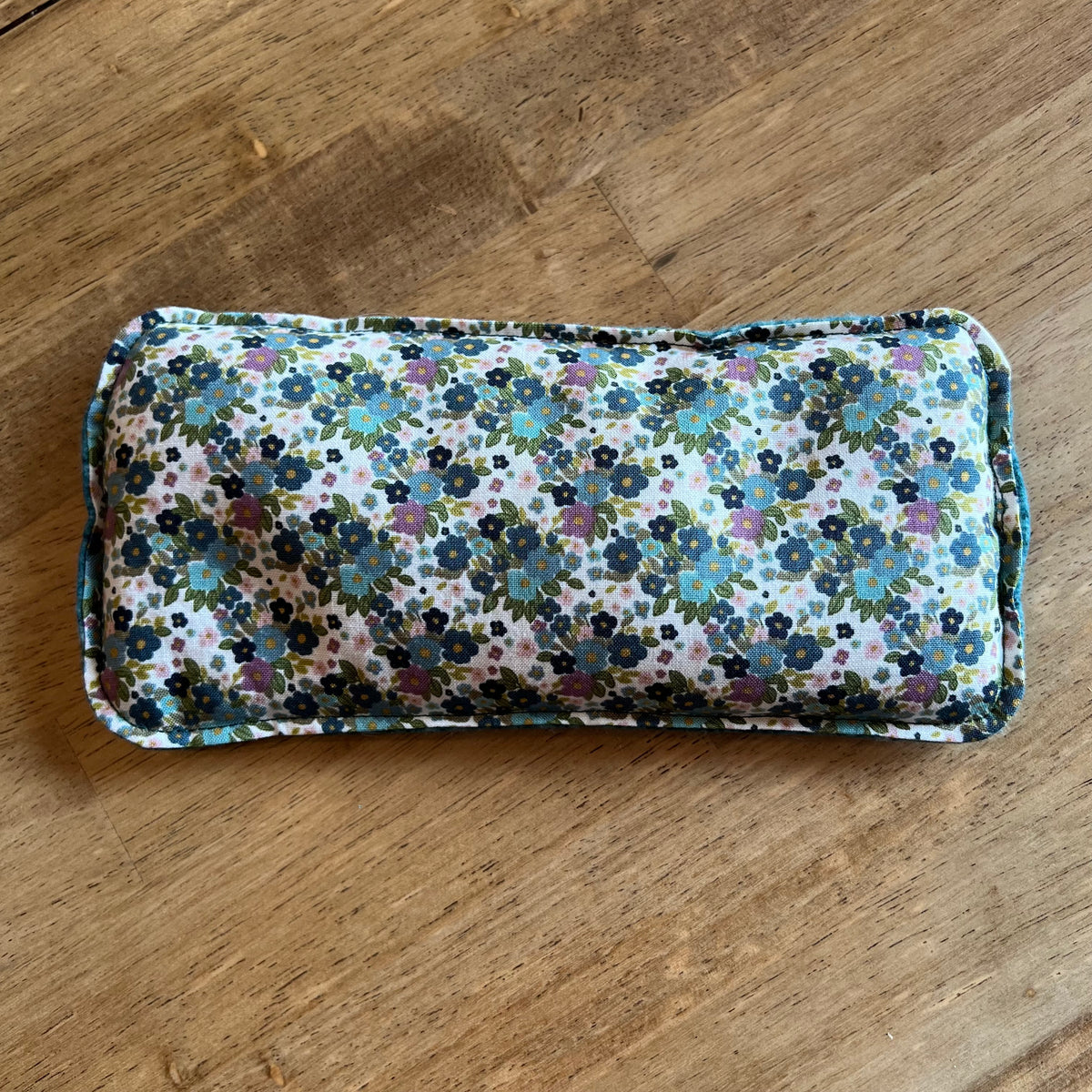 Dual-sided Wildflower Glen Weighted Eye Pillow by Eclectic Spruce. This pillow features white fabric containing a colorful floral print in shades of blue, green, pink, and purple.