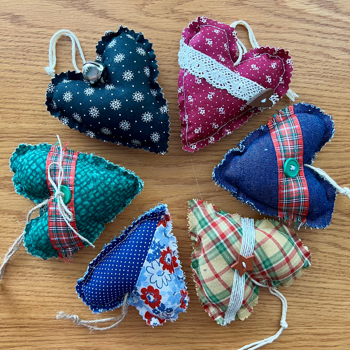 Eclectic Spruce's handmade fabric heart-shaped ornaments are made of cotton, stuffed with fiberfill, and embellished with lace, buttons, bells, and other trim.  Sold mystery style, you will receive a unique heart (or curated combination of hearts) picked out just for  you. This photo shows 6 Eclectic Spruce heart ornaments arranged in a circle on a wooden surface.