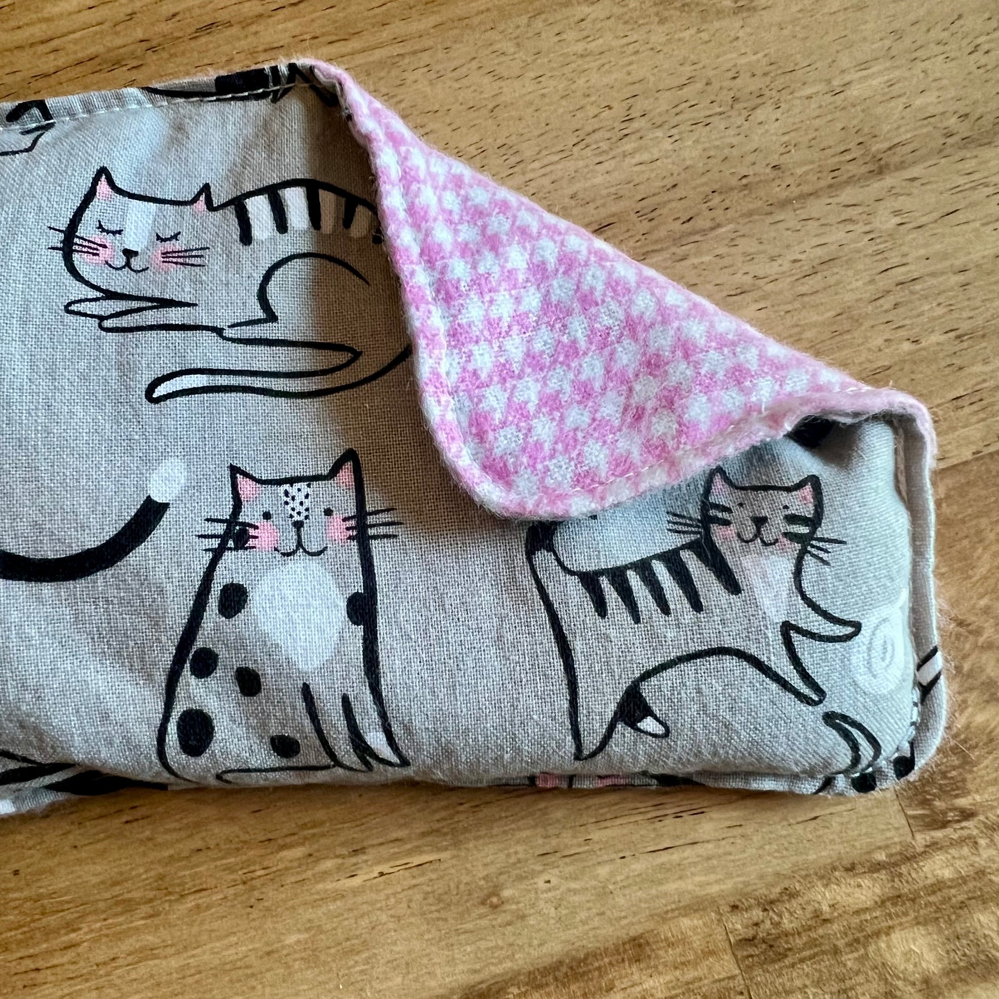 A close-up of the Purrfect Pause Weighted Eye Pillow by Eclectic Spruce. The front side is a light grey cotton fabric with black, white, and pink illustrated cats. The corner of the pillow is folded over, revealing a light pink and white houndstooth-patterned flannel fabric on the reverse side.
