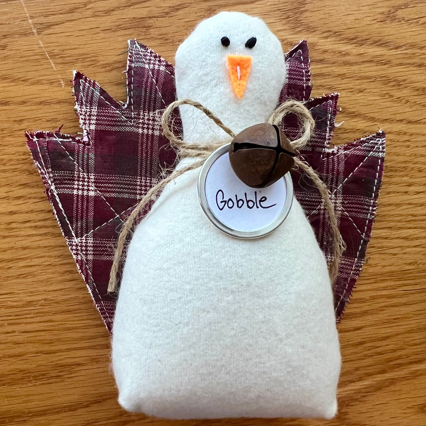 An Eclectic Spruce handmade fabric Thanksgiving turkey sitting on a wooden table. The turkey features a white flannel body, textile tail feathers, an orange felt beak, black stitched eyes, a brown rusty bell on a twine bow, and a round white tag that reads "Gobble."
