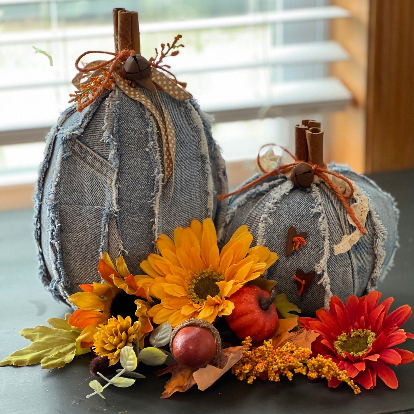 Two Eclectic Spruce decorative pumpkins made from faded blue denim fabric with rustic fringe, faux cinnamon sticks, ribbon, and twine. They are styled with autumn flowers and leaves on a dark tabletop in front of a window.
